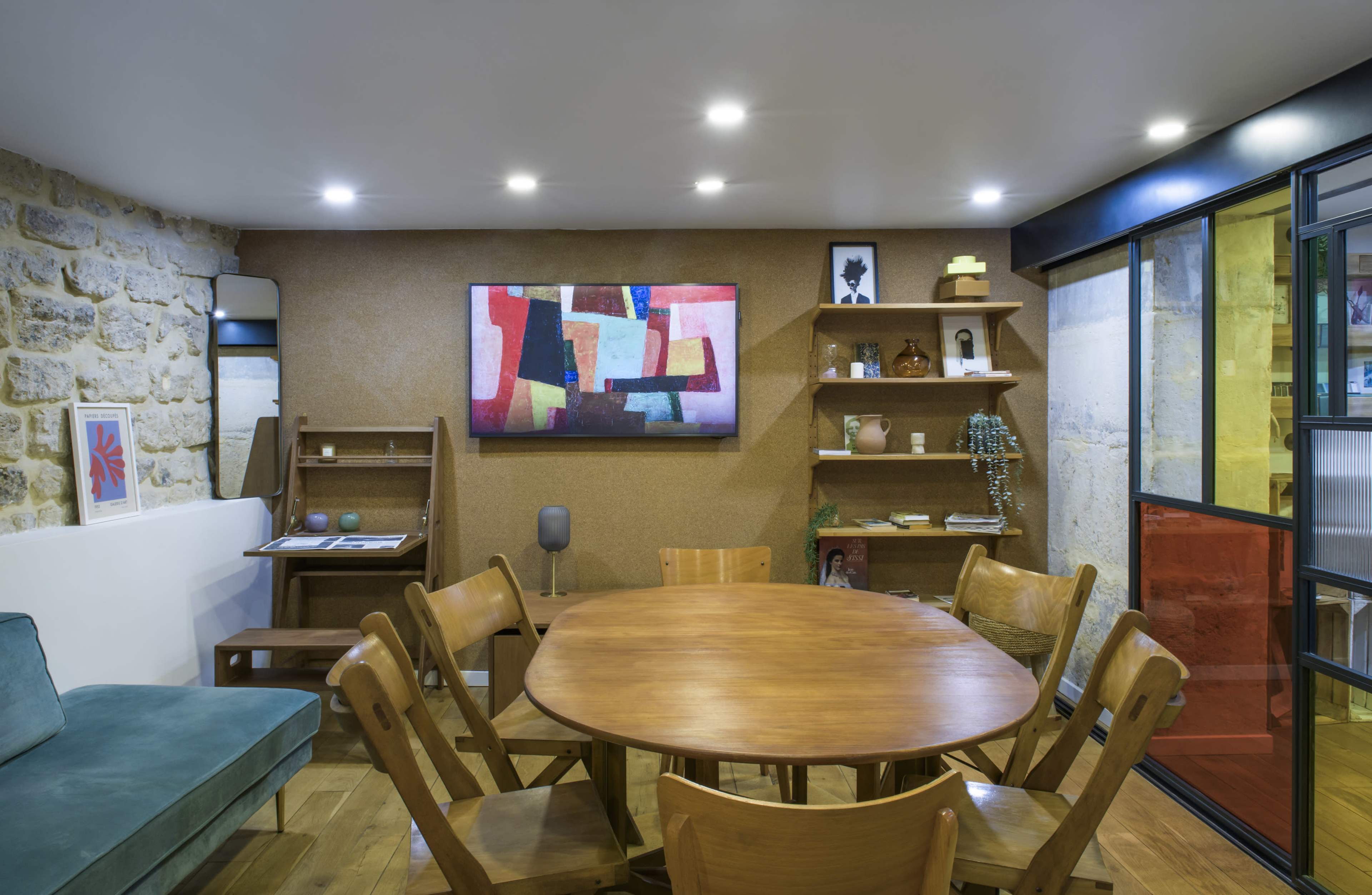 The image shows a modern interior space featuring a round wooden dining table surrounded by wooden chairs, a couch, and shelves displaying various decorative items.