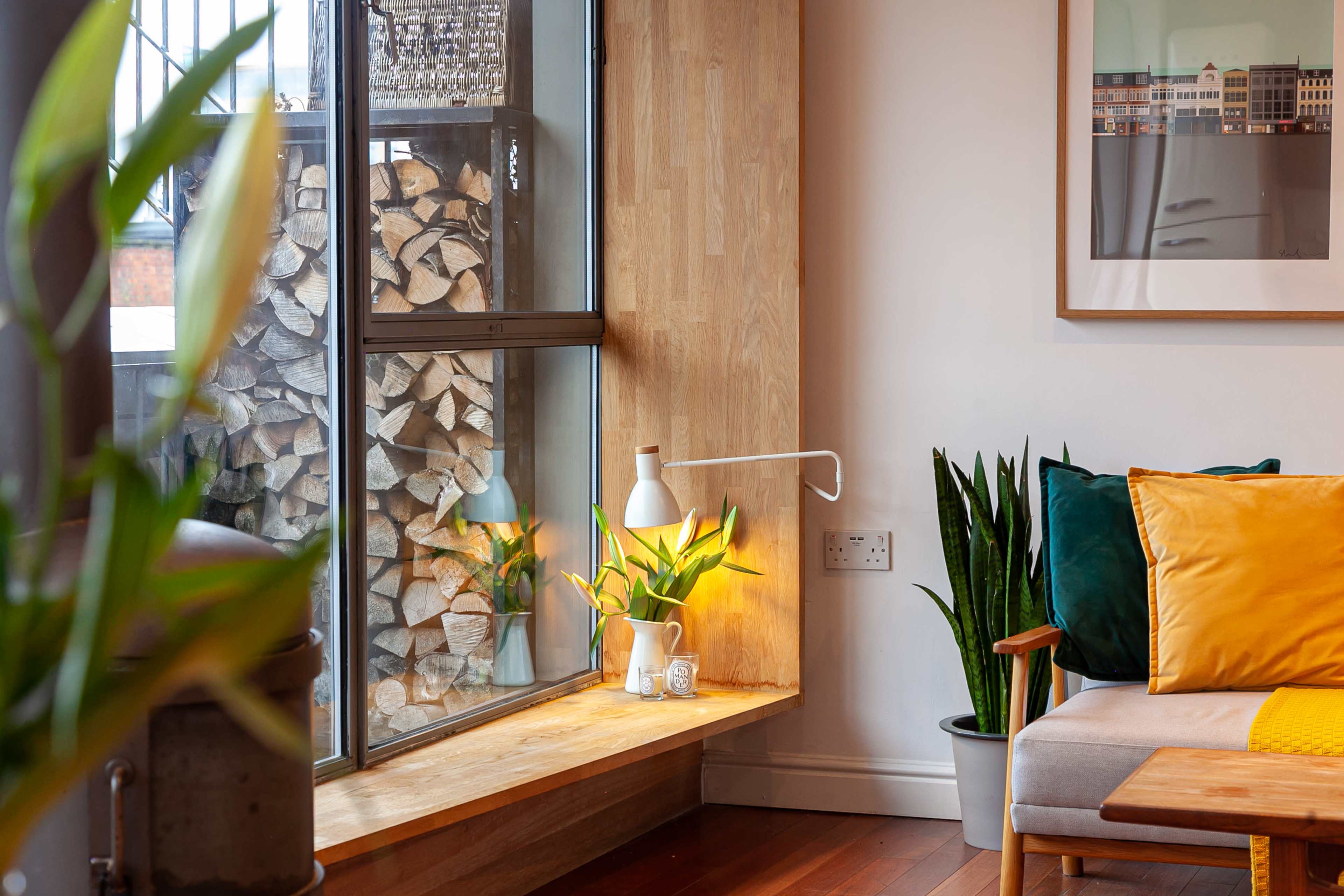 A well-lit corner of a living room features a wooden window sill with plants, a lamp, and a stack of logs visible through the glass.