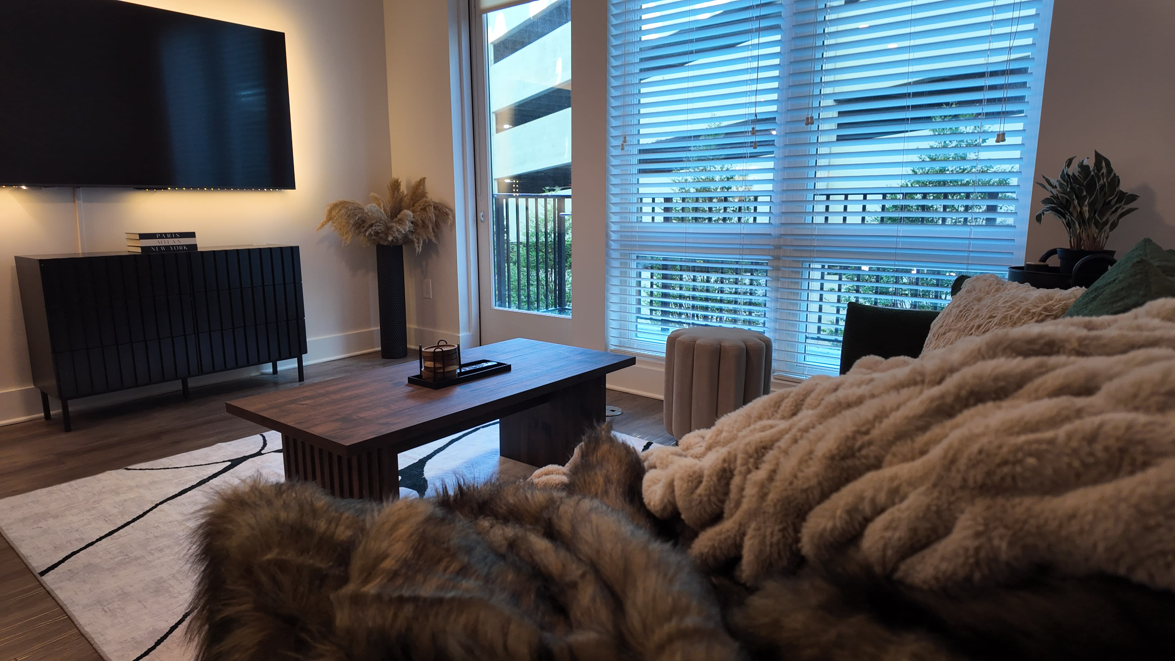The image shows a modern living room featuring a wooden coffee table, a black entertainment unit, a large television, and large windows with blinds letting in natural light.