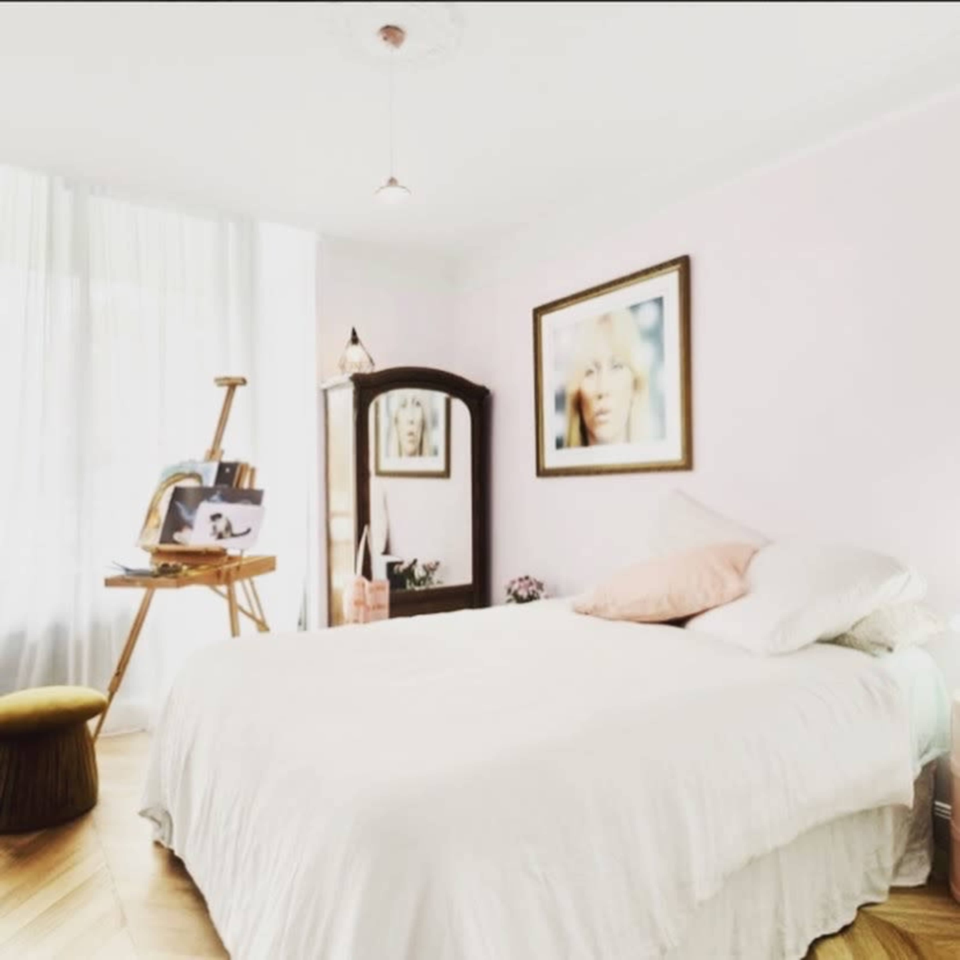 A light-colored bedroom features a bed with white linens, a small stool, an easel displaying art, a mirror, and a framed portrait on the wall.