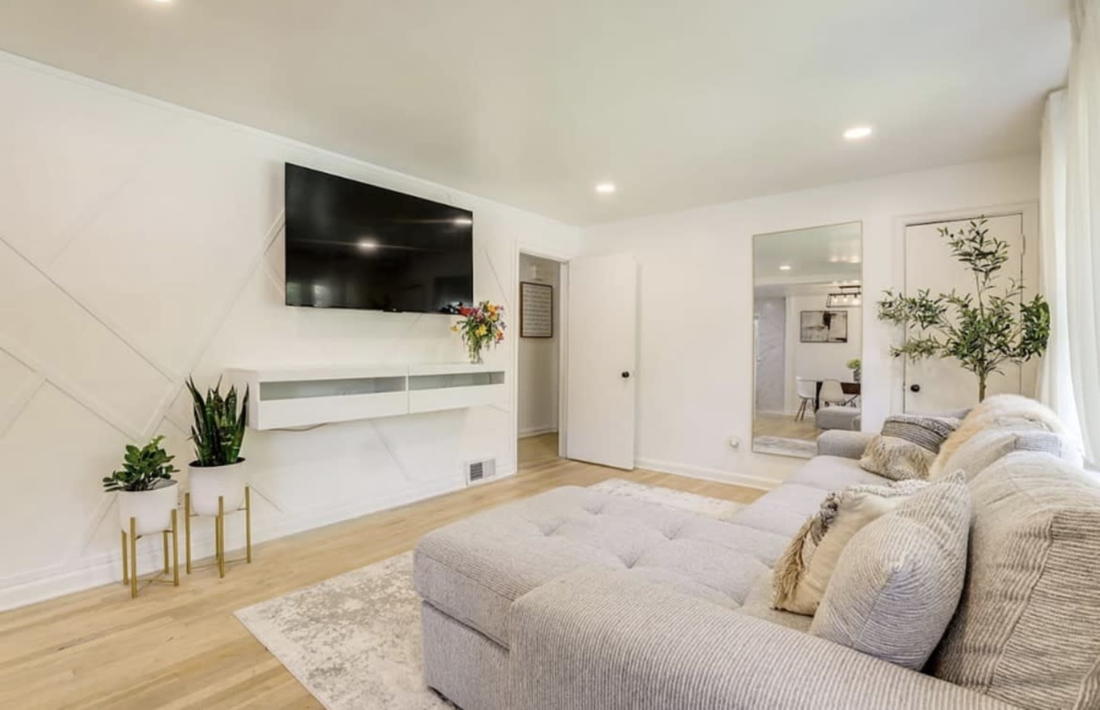A modern living room features a large sectional sofa, a wall-mounted television, and a minimalistic decor with plants and mirrors.