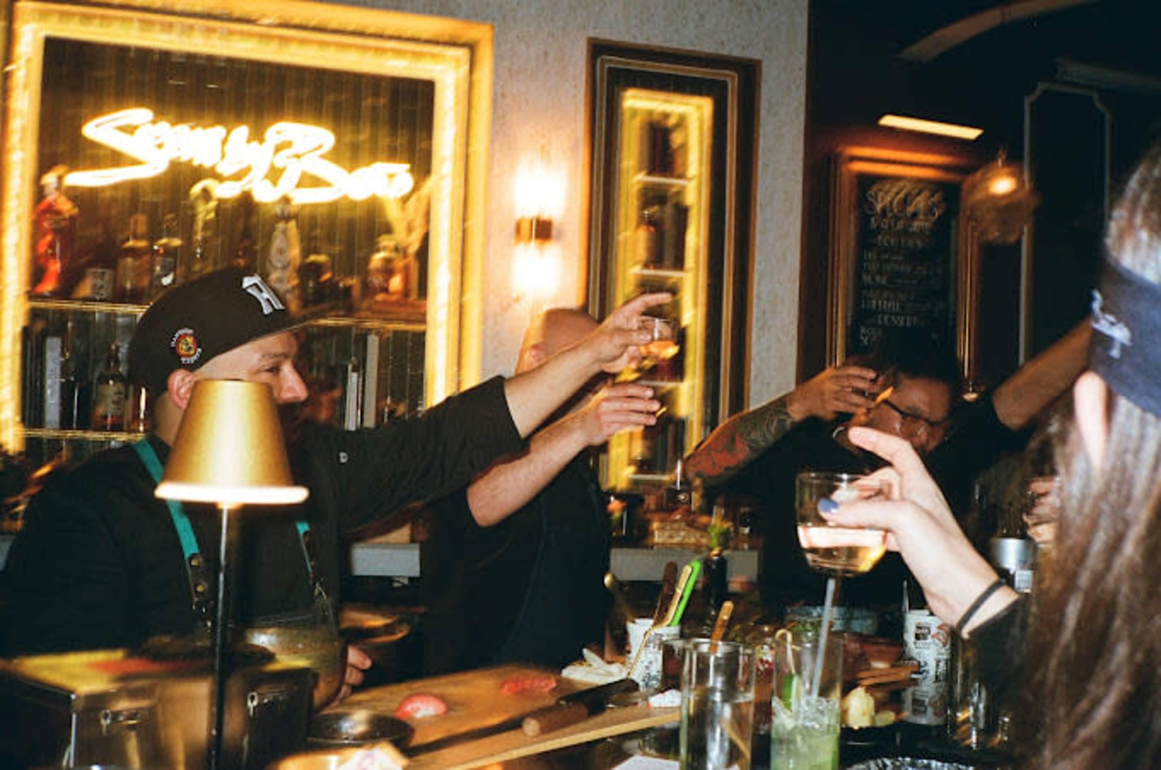 A group of people raise their glasses in a toast at a bar, with a neon sign in the background.