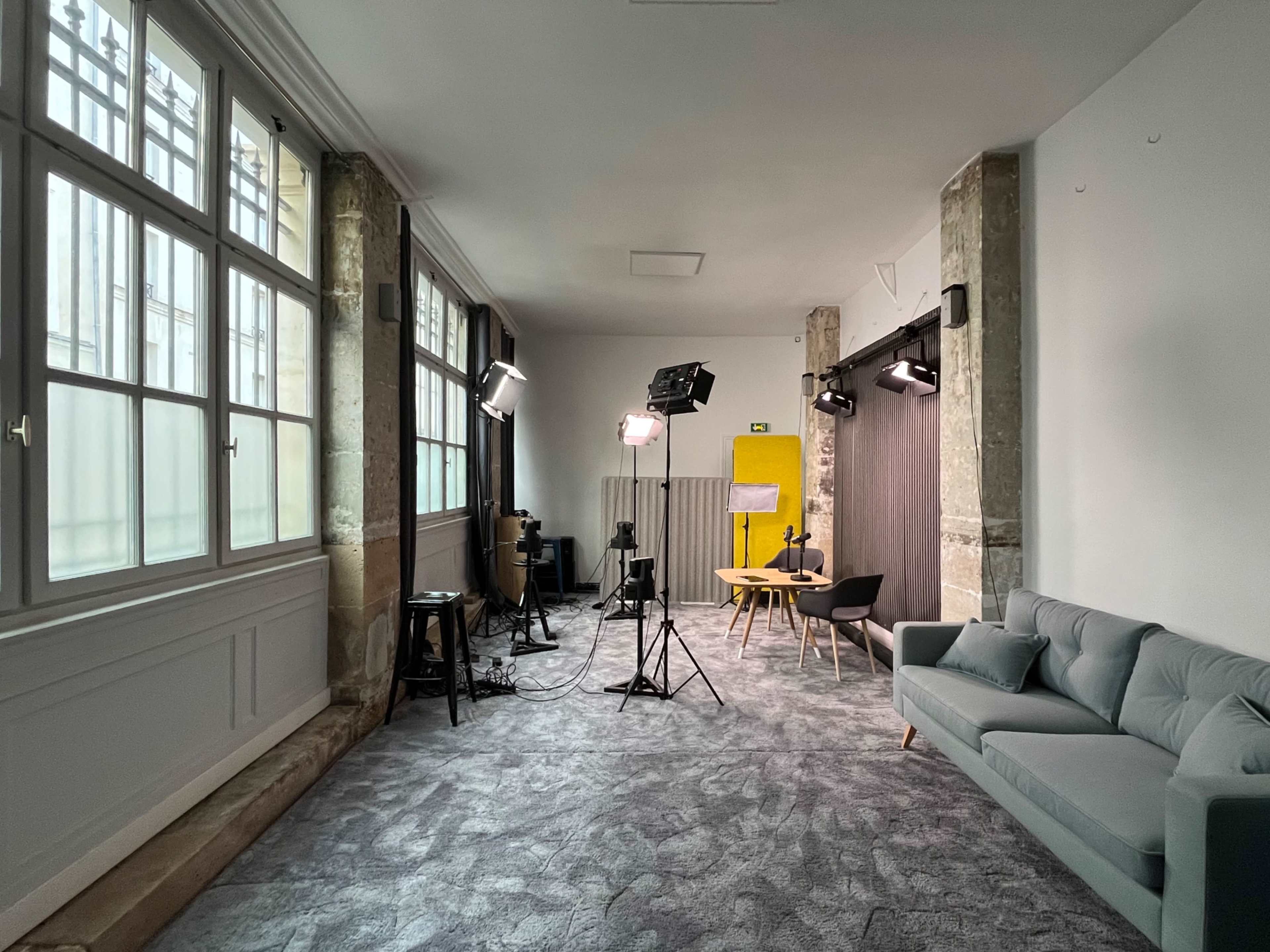The image shows a well-lit studio space featuring a gray sofa, a wooden table with chairs, and professional lighting equipment set up against a backdrop.