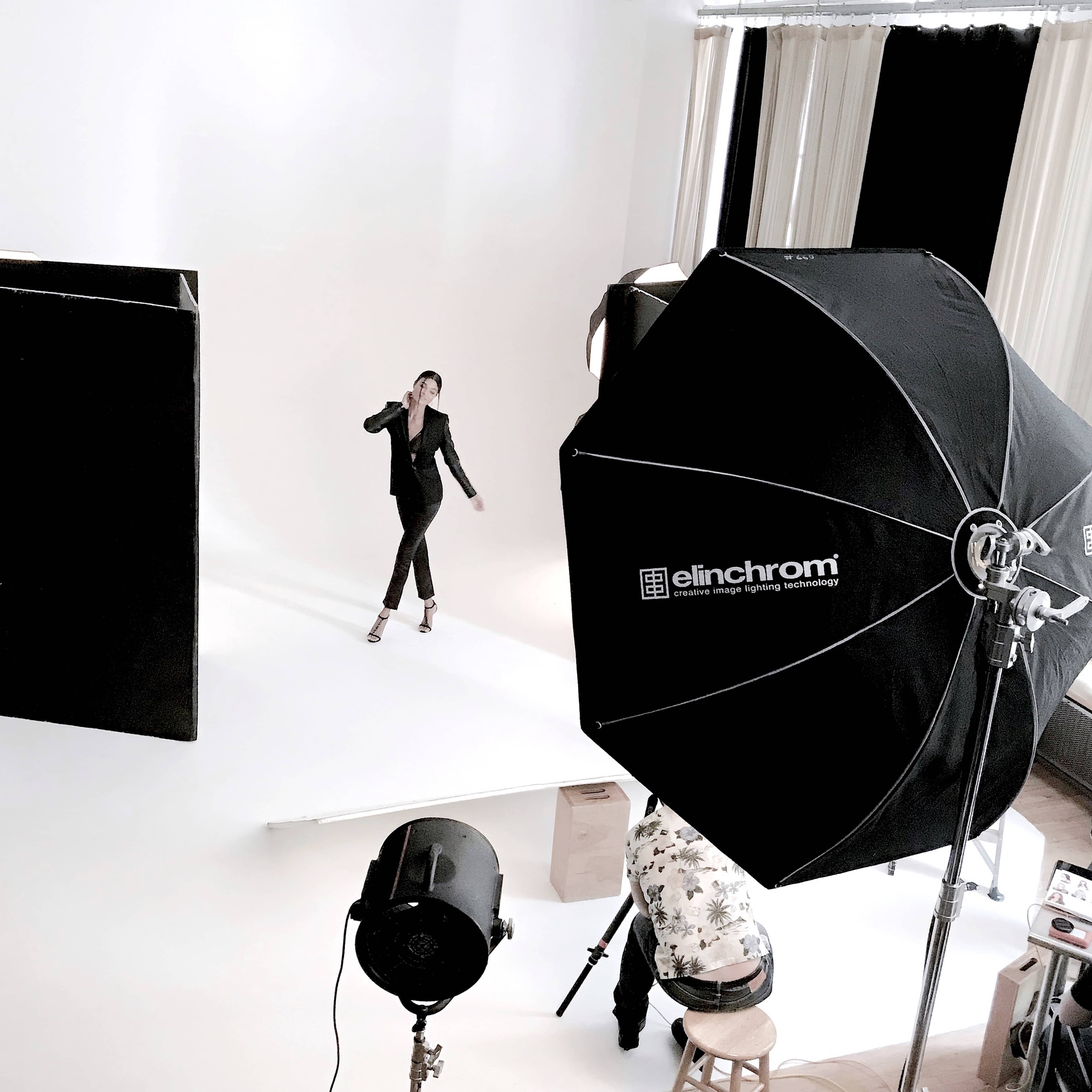 A model poses on a white backdrop during a photoshoot, while a large softbox and studio lights illuminate the scene.