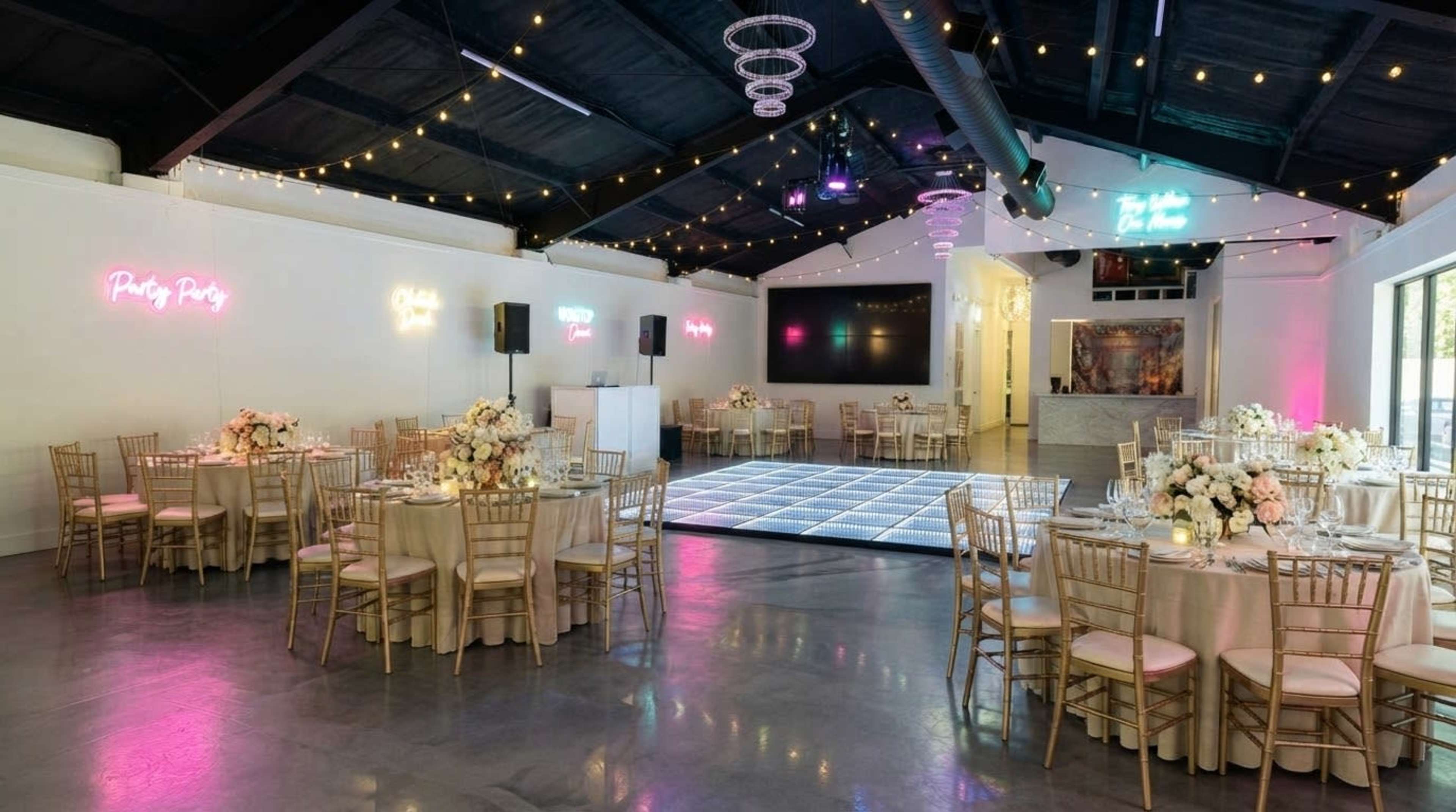 The image shows an elegantly decorated event space featuring circular tables with floral centerpieces, a dance floor, and neon sign displays.
