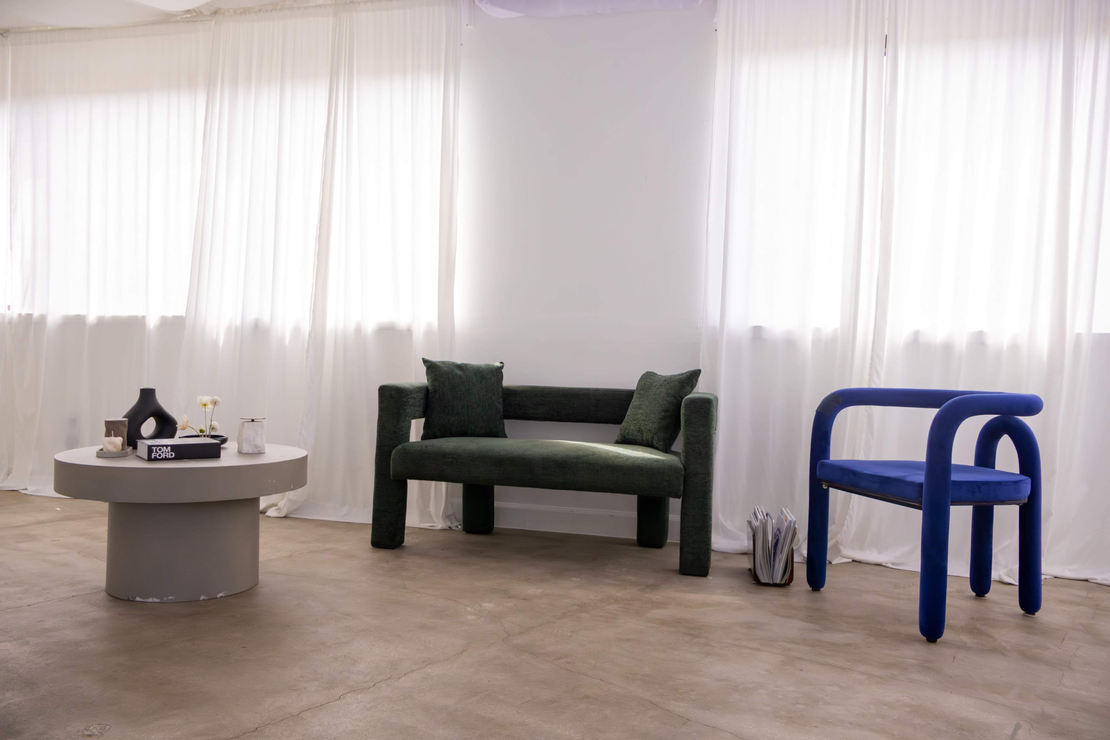 The image shows a modern interior space featuring a green upholstered sofa, a round gray coffee table, and a blue armchair against a backdrop of sheer white curtains.