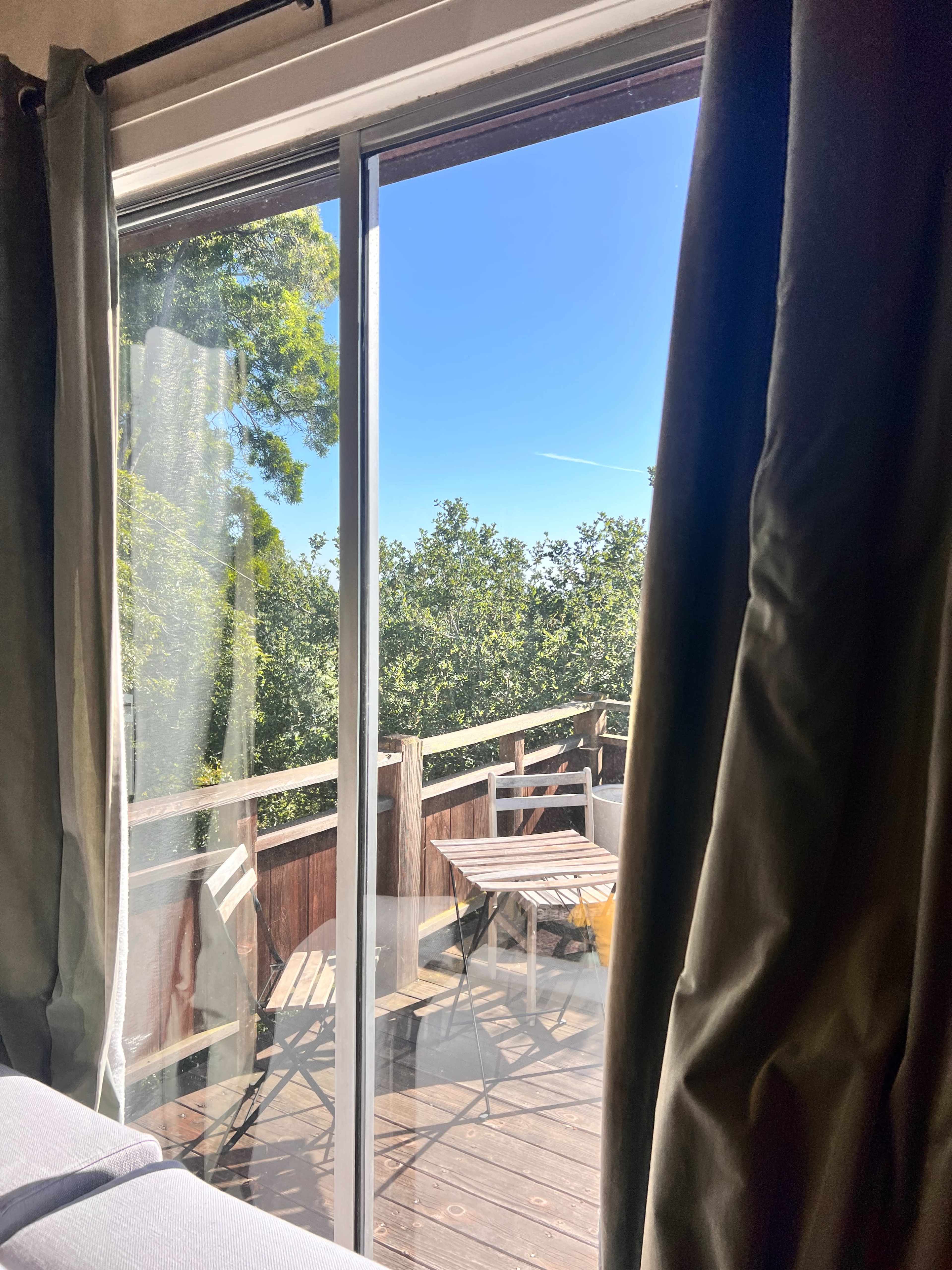 A window with sheer curtains frames a view of a balcony with a small table and chairs, surrounded by trees under a clear blue sky.