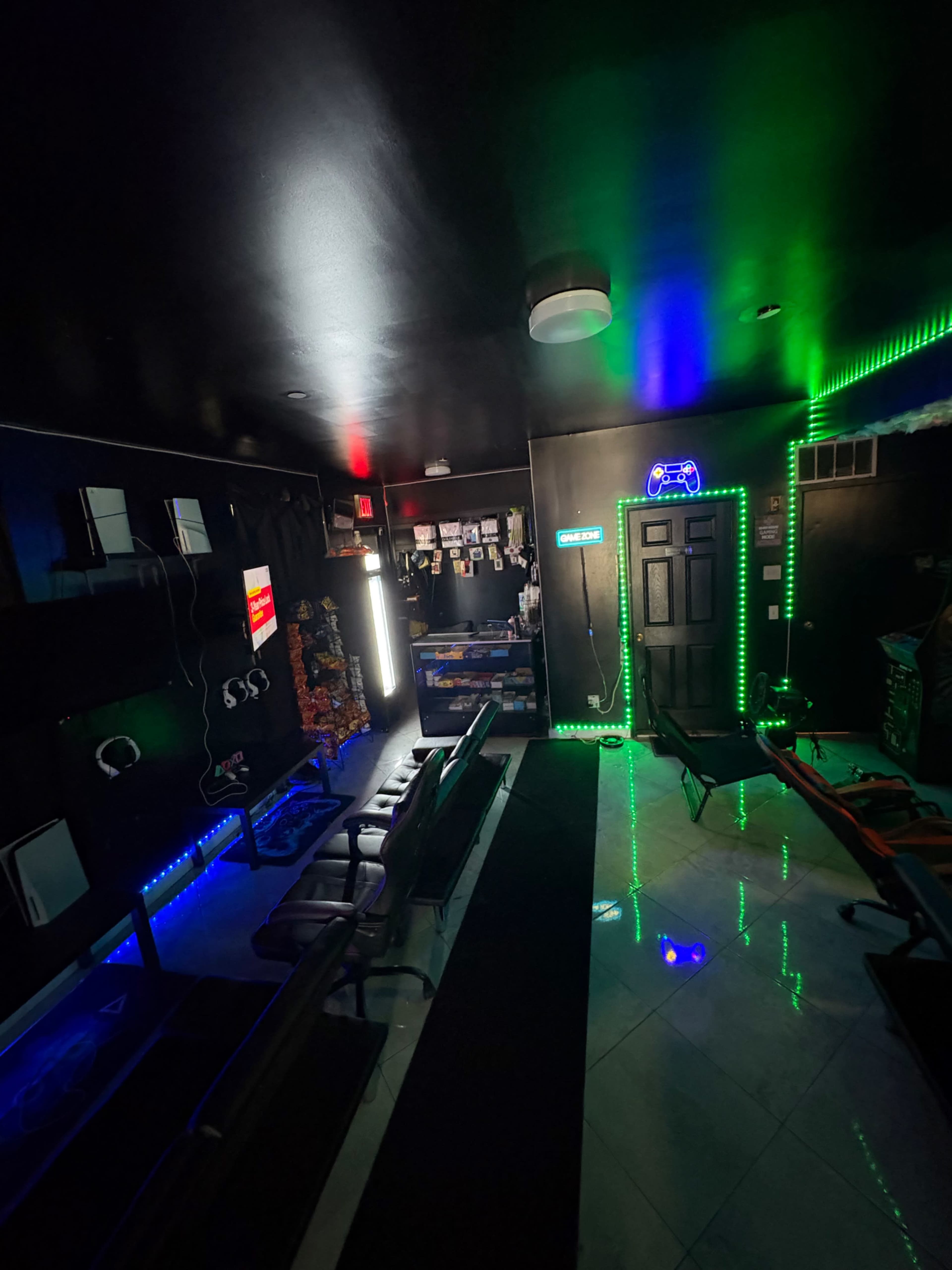 The image shows a dimly lit room with black walls, colorful lights, and multiple seating areas, likely designed for gaming or entertainment activities.