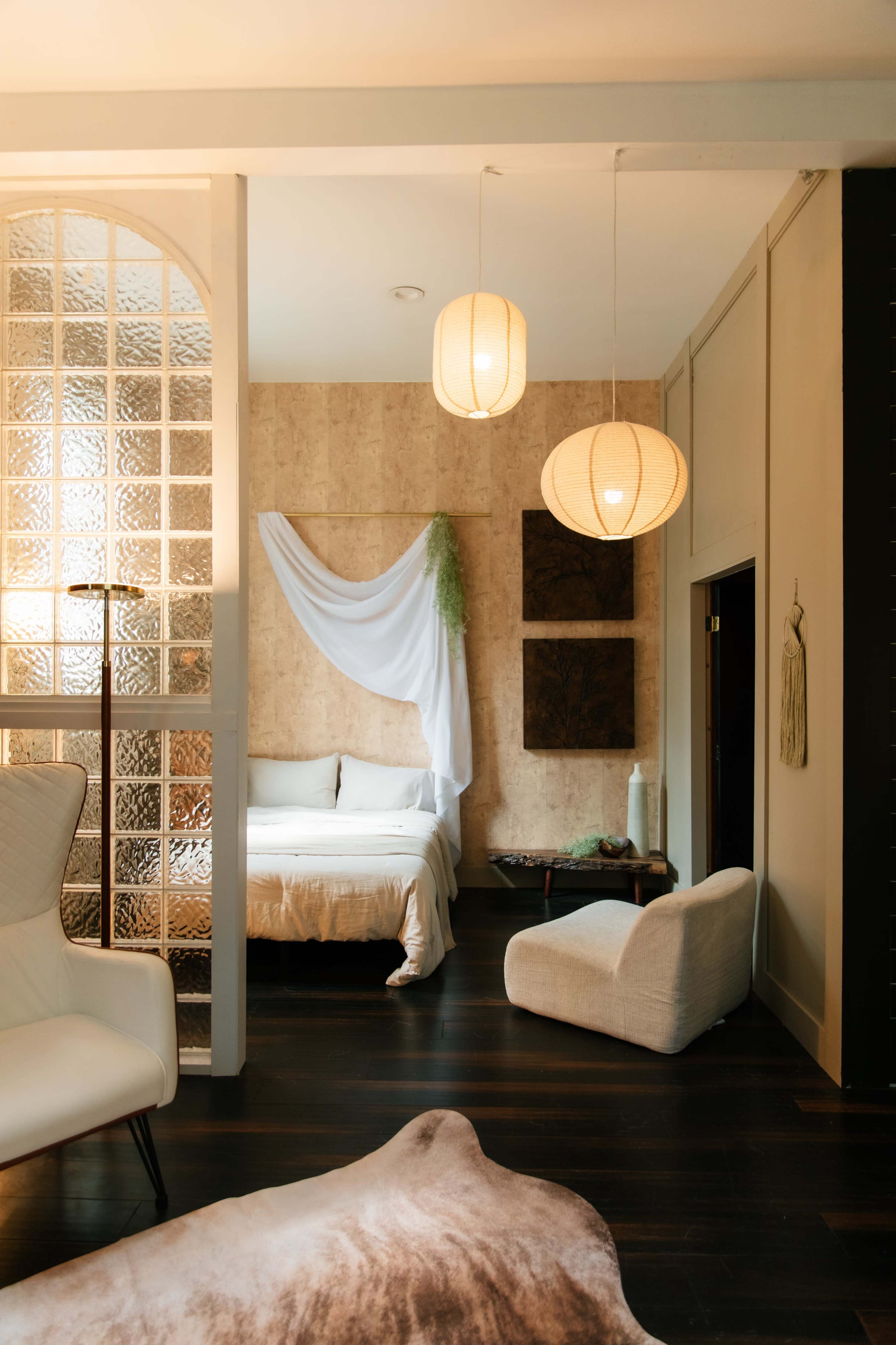 The image shows a softly lit bedroom featuring a bed with white linens, decorative wall hangings, and two round pendant lamps.