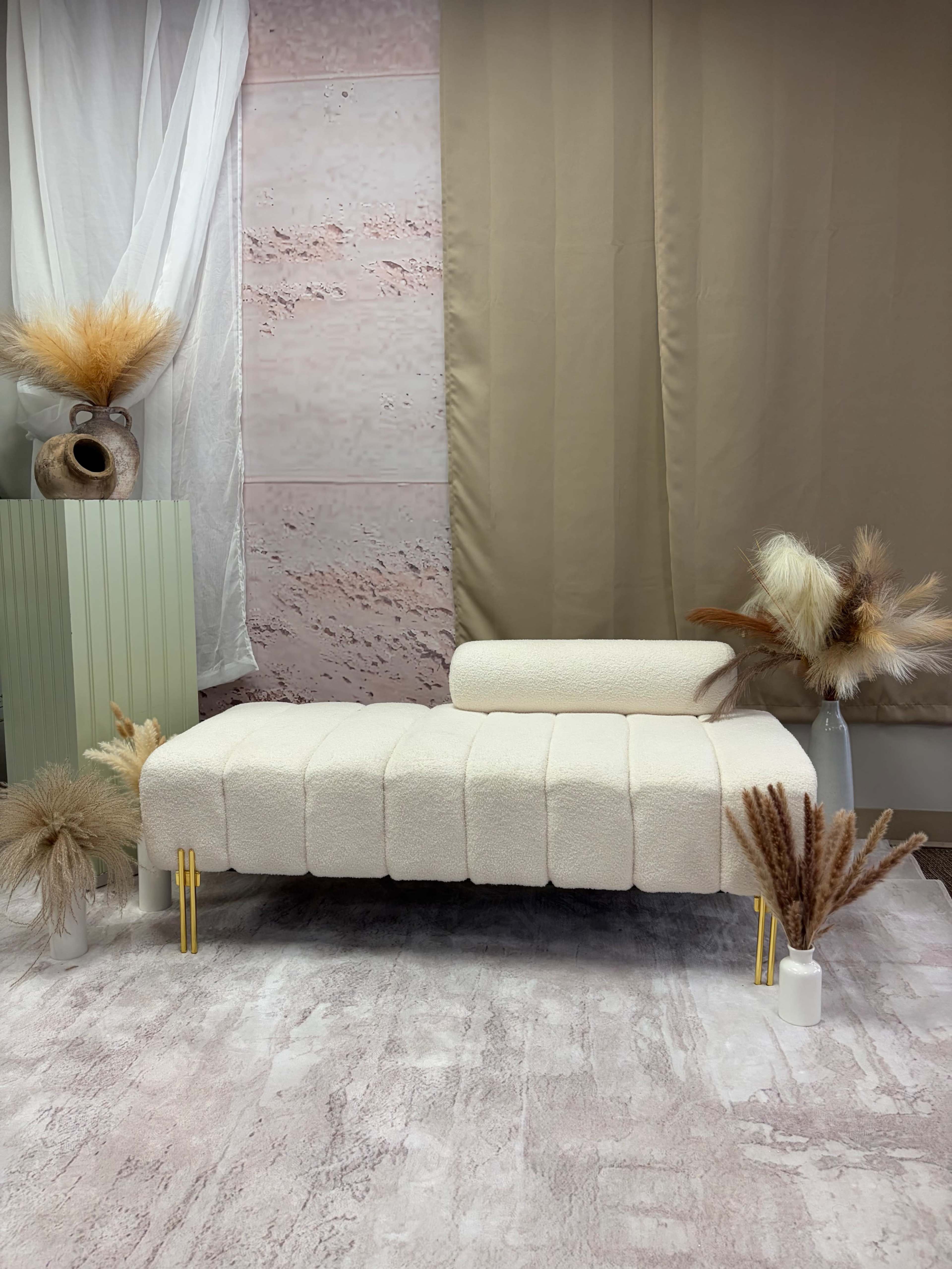 The image features a modern, cushioned bench with a removable backrest, surrounded by beige and cream decorative elements, including dried plants and a patterned background.