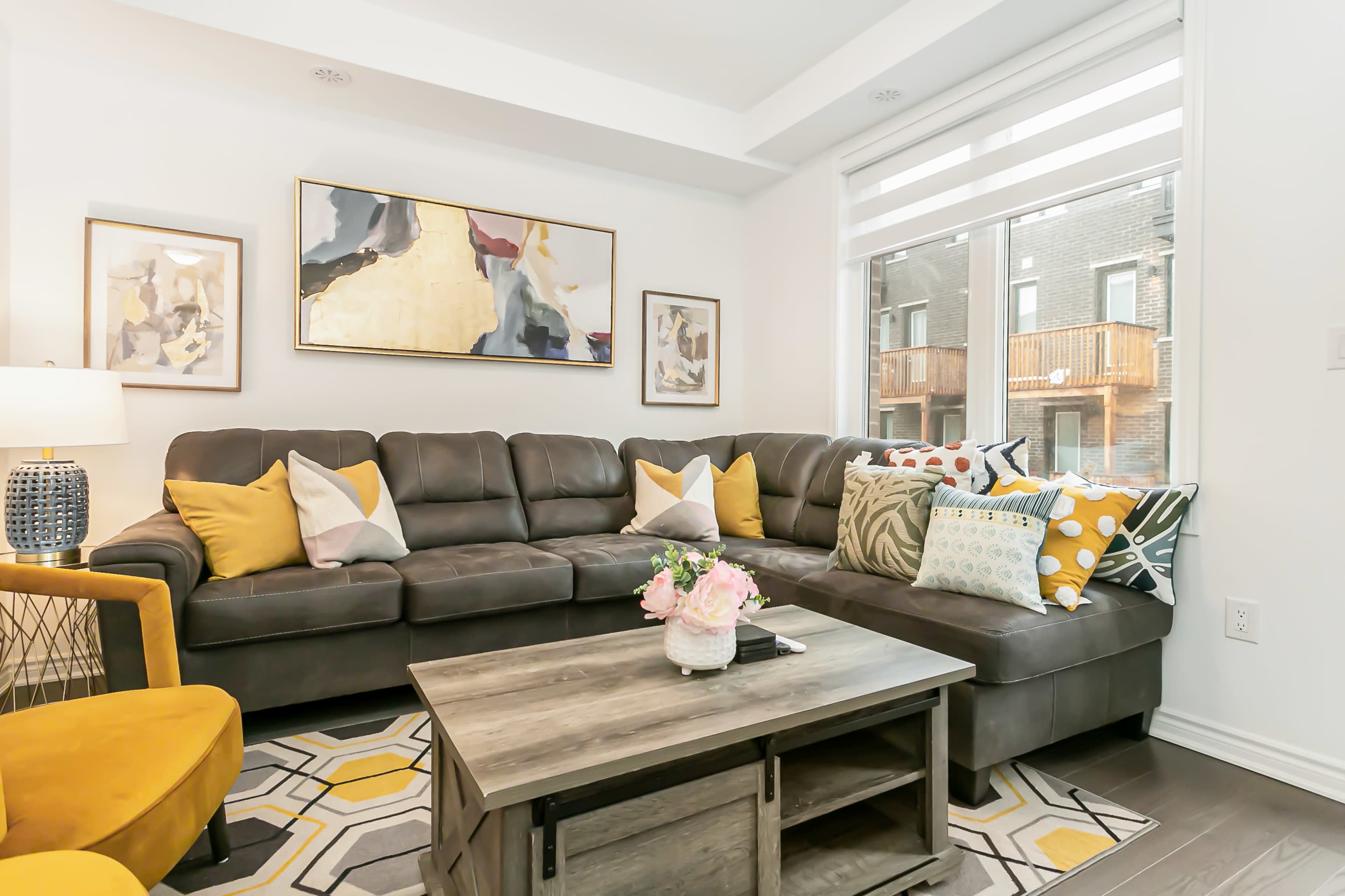 A modern living room features a sectional couch with colorful cushions, a wooden coffee table, and large windows showcasing an outdoor balcony.