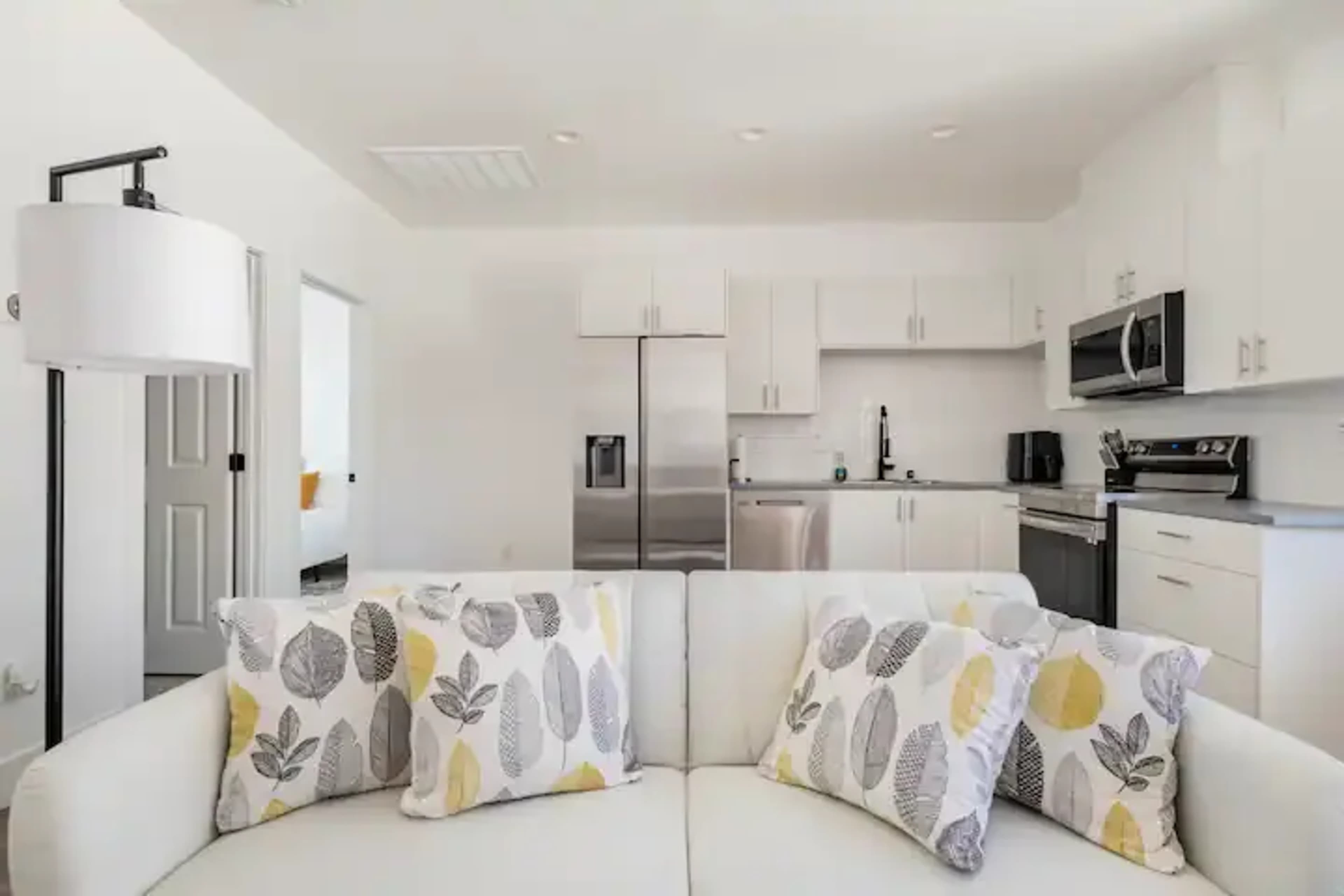 A modern kitchen and living area are shown, featuring a white sofa with decorative pillows and stainless steel appliances in the background.