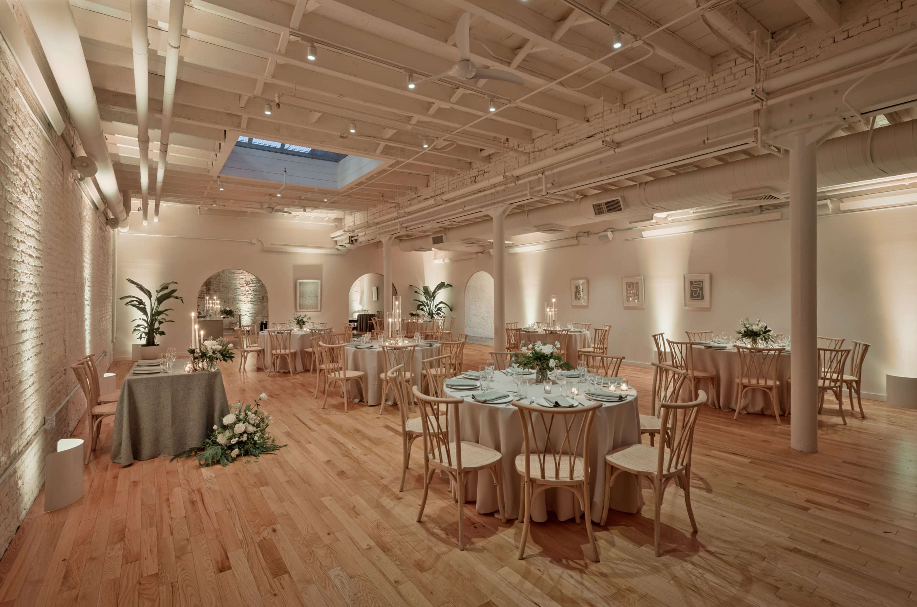 A spacious, elegantly arranged event venue with round tables set for dining, surrounded by light wood flooring and soft lighting.