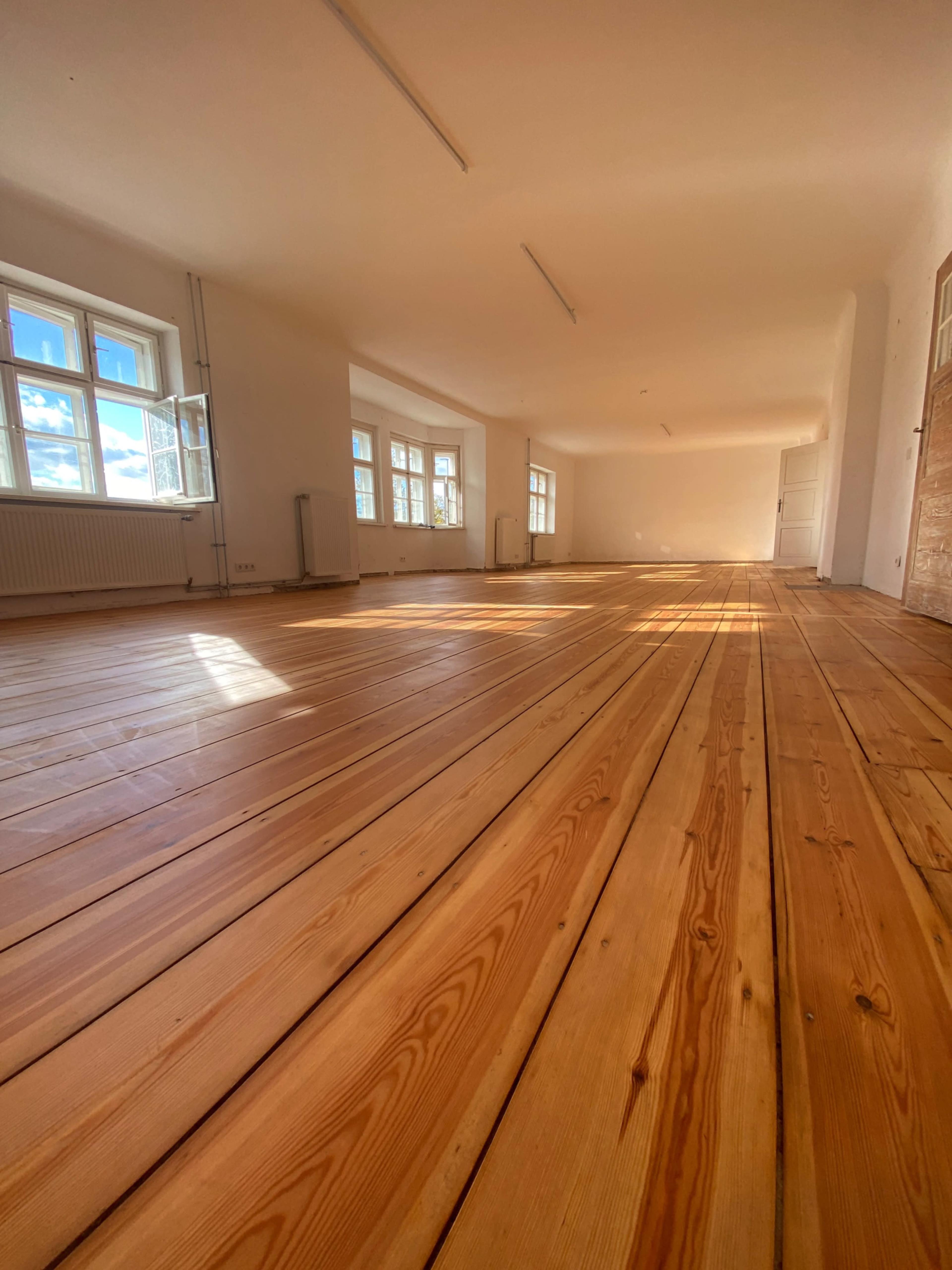 The image shows a spacious, empty room with wooden flooring and large windows allowing natural light to enter.