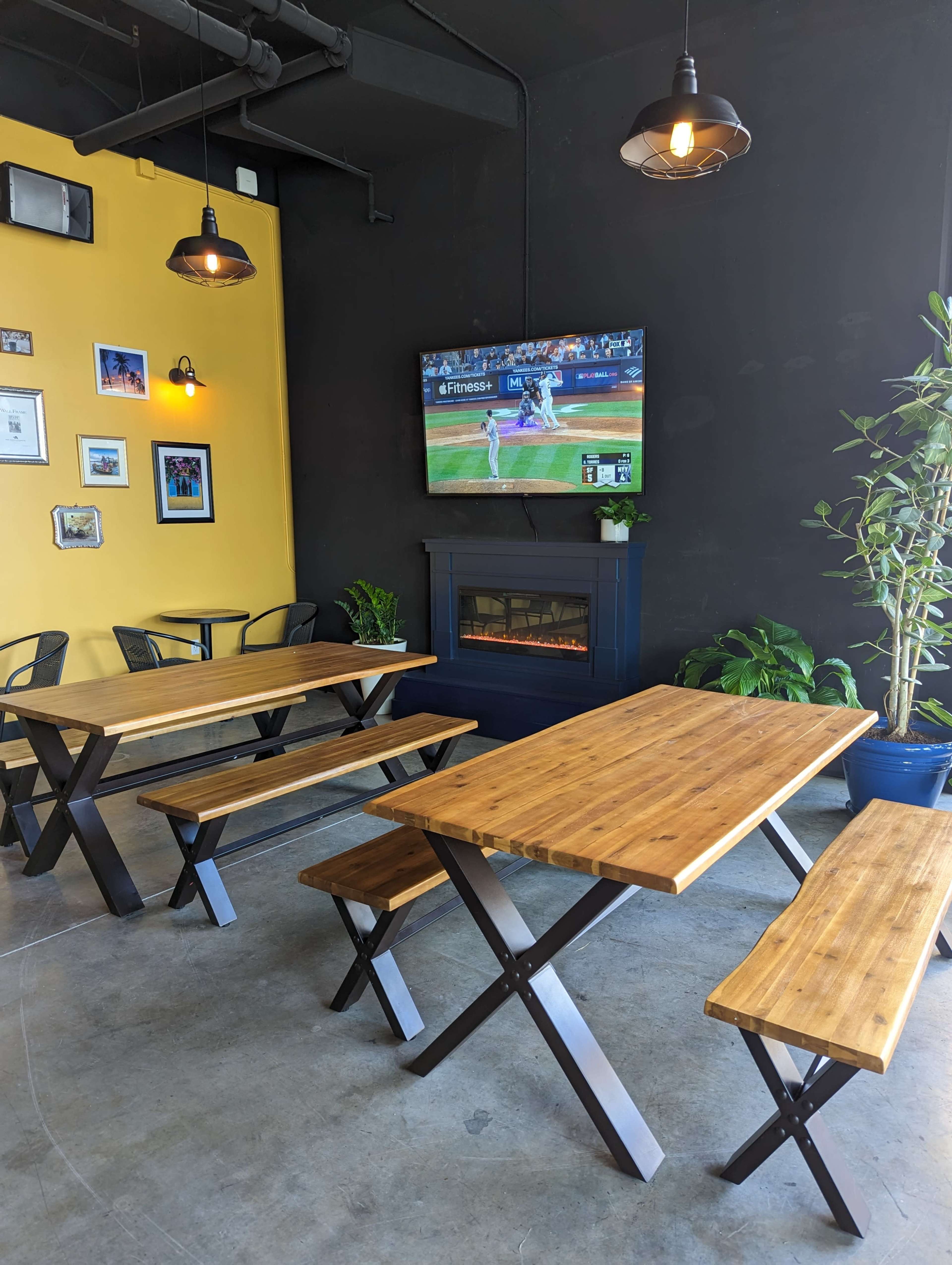 A modern dining area features wooden tables and benches, a television displaying a sporting event, and a small fireplace against a contrasting yellow and black wall.