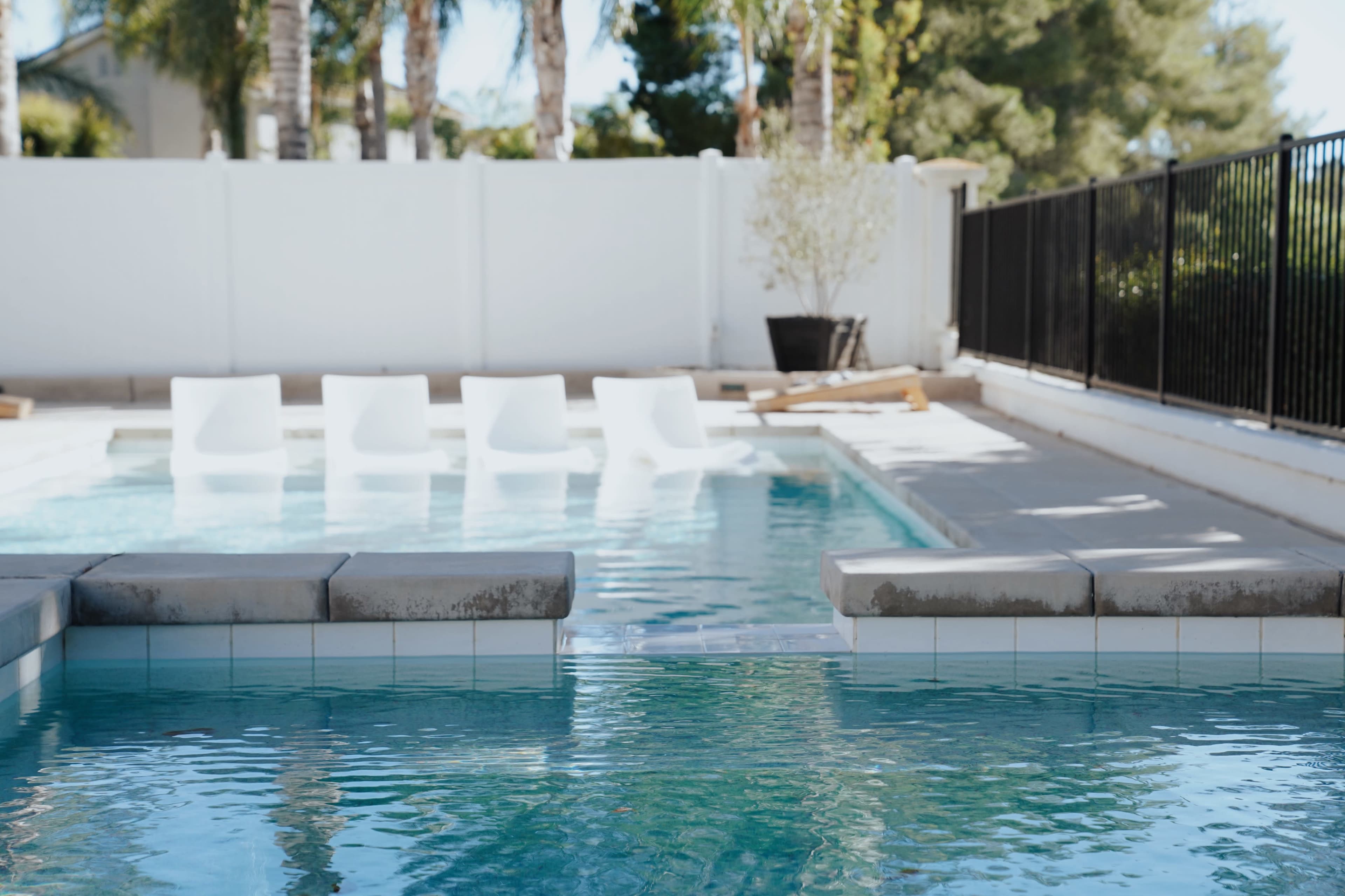 A clear swimming pool with white lounge chairs positioned along its edge and a fenced yard in the background.
