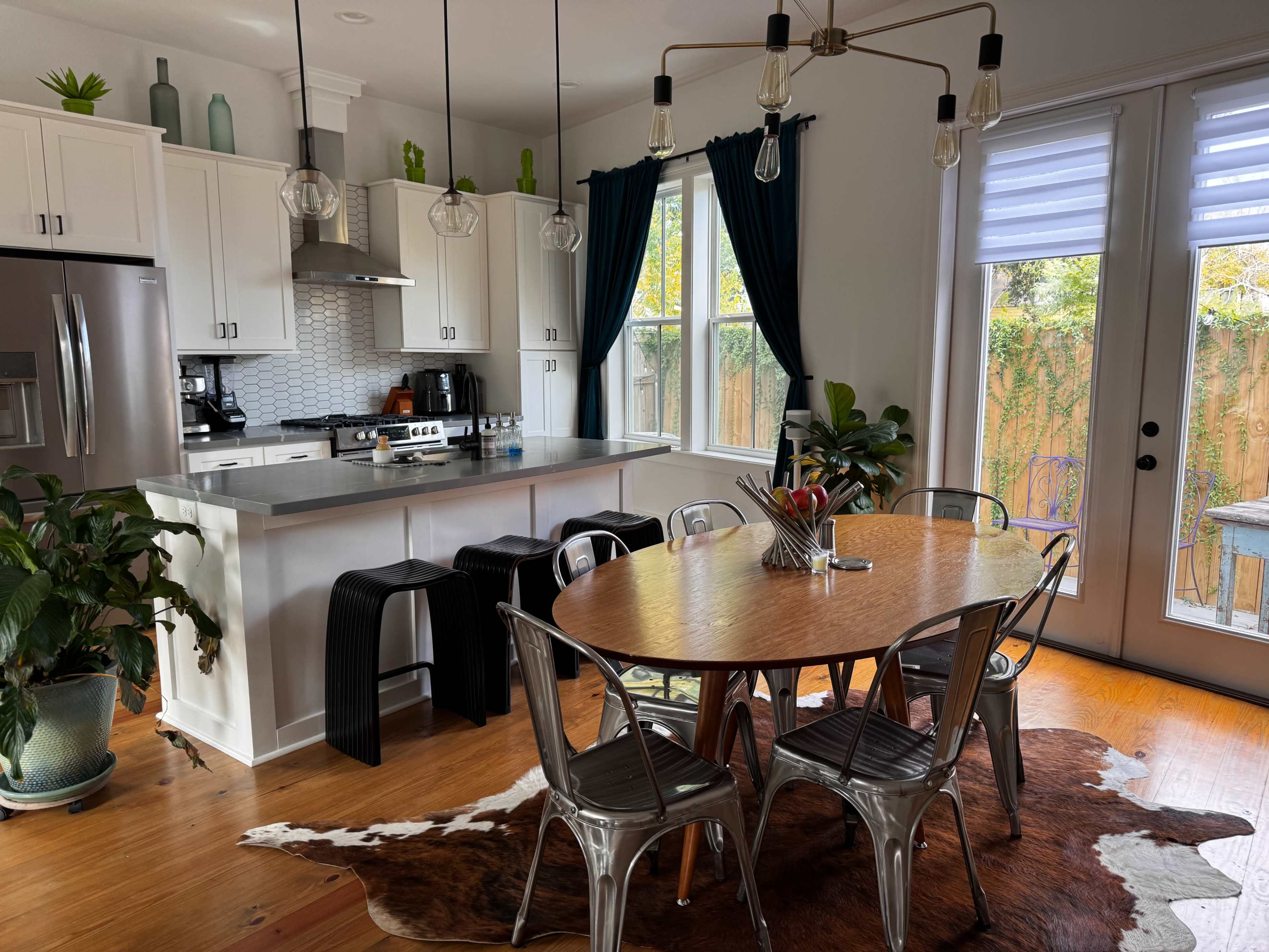 A modern kitchen and dining area with a circular wooden table surrounded by metal chairs, a countertop with bar stools, and large windows letting in natural light.