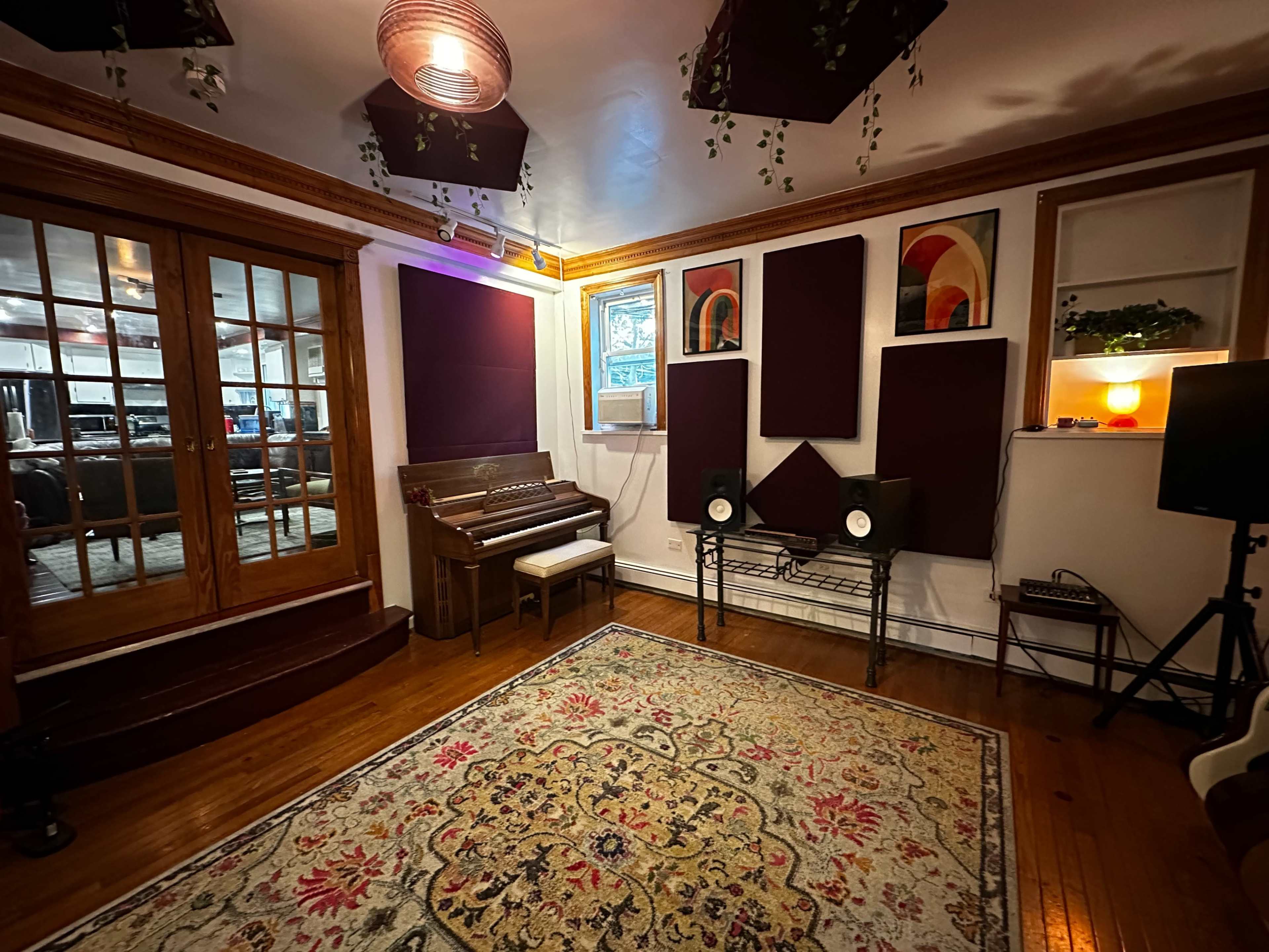 The image shows a cozy music studio featuring a piano, acoustic panels on the walls, a decorative rug, and a pair of speakers positioned on a shelf.