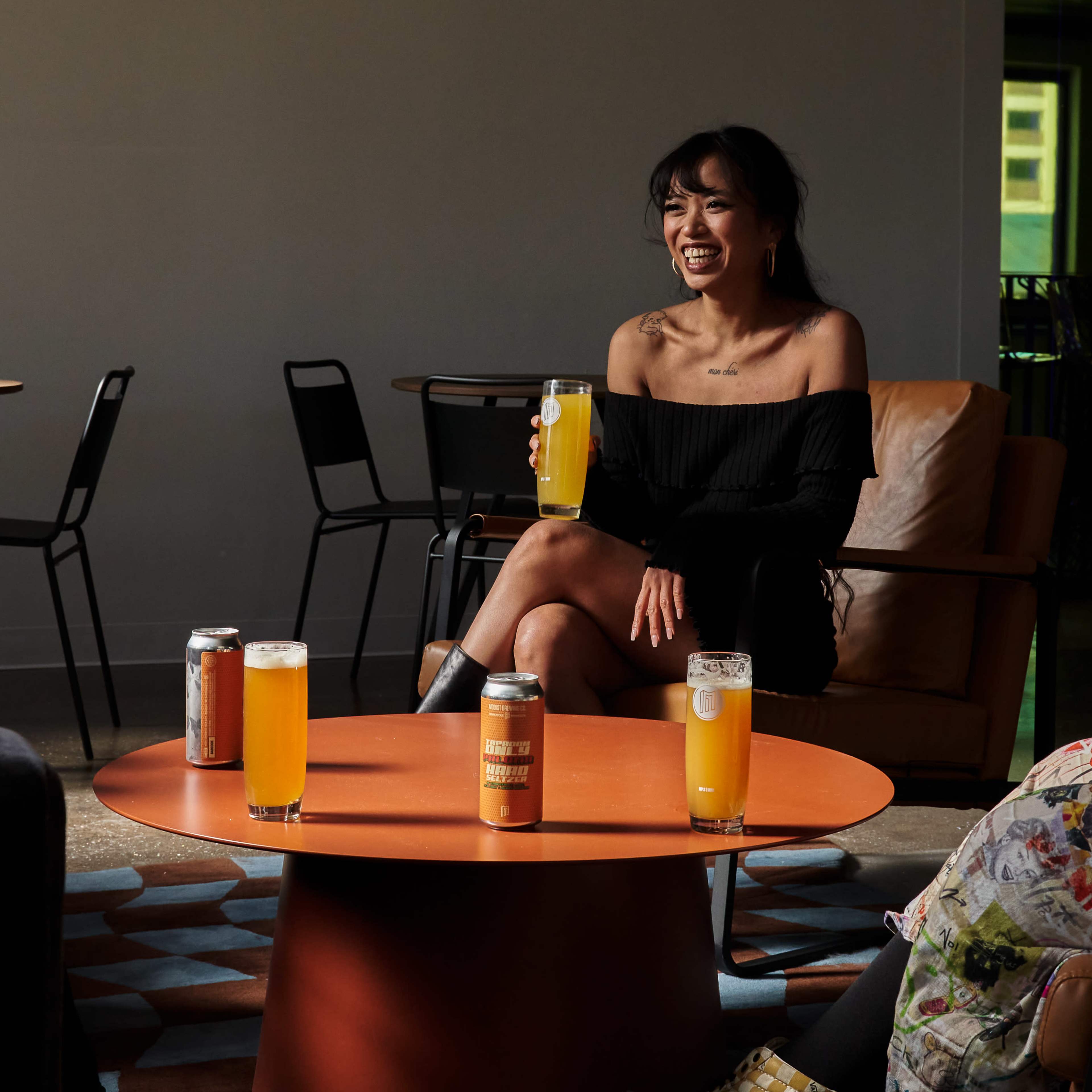 A woman sits on a chair holding a drink, surrounded by several cans and glasses of beer on an orange table in a relaxed setting.