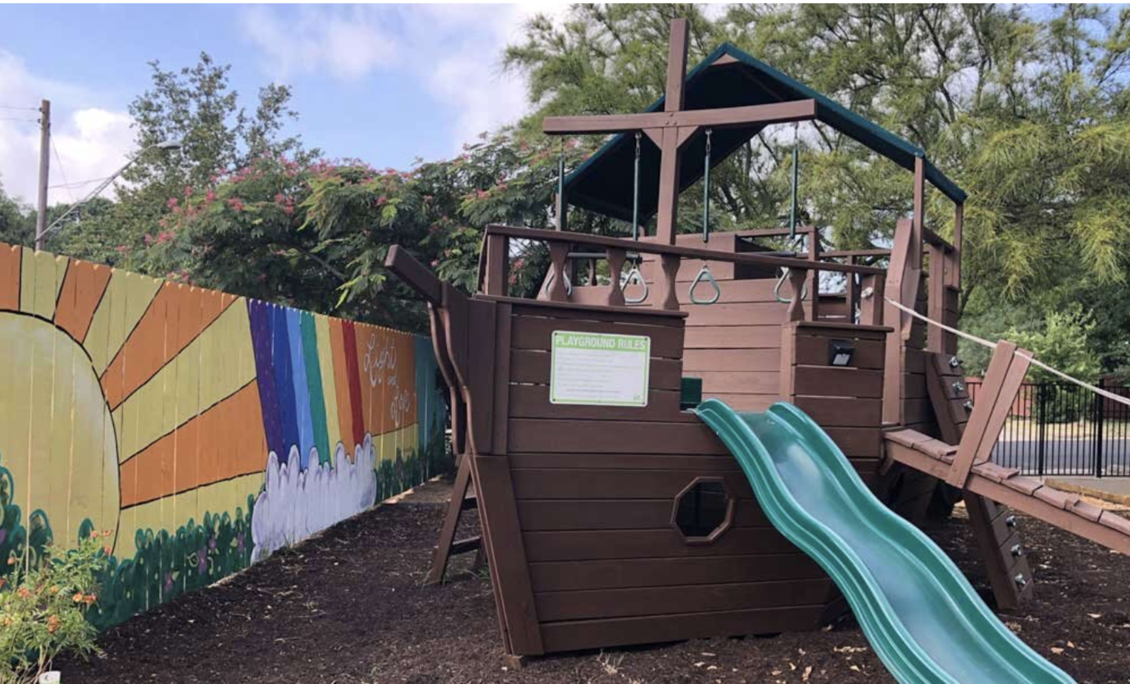 A wooden playground structure designed like a ship features a green slide and climbing accessories beside a colorful mural.