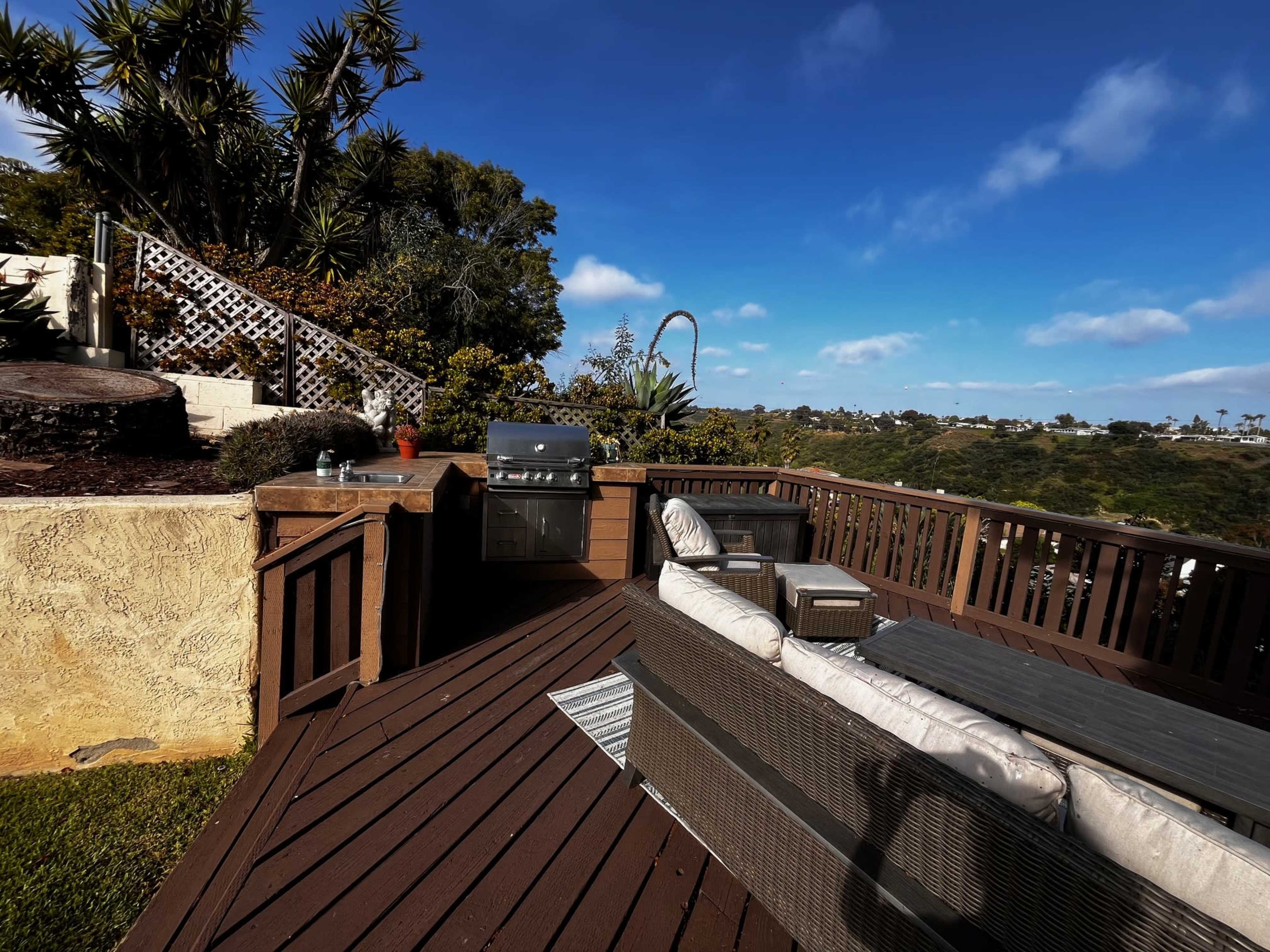 A wooden deck features a grill and outdoor seating, overlooking a green hillside under a blue sky.