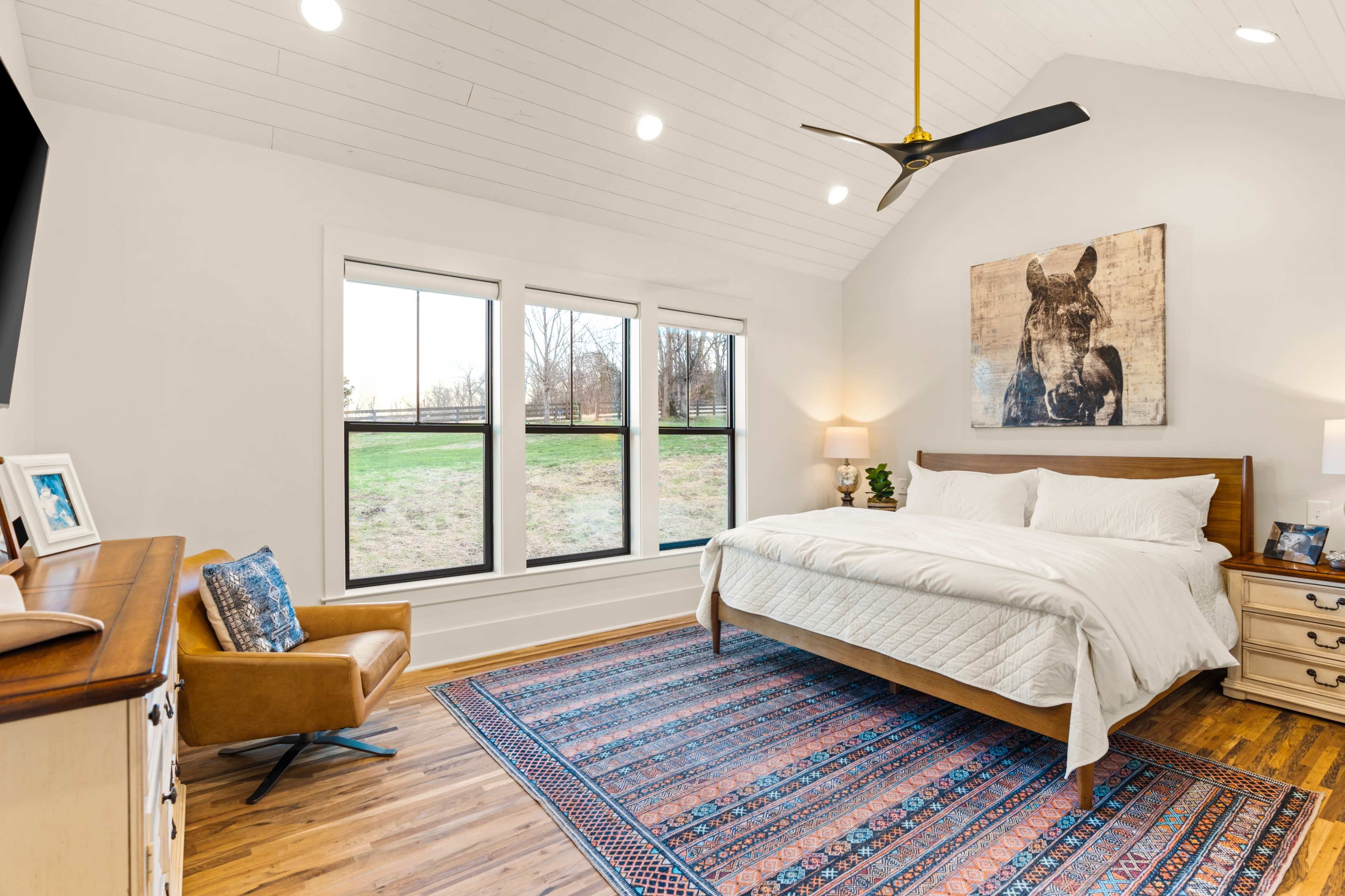 The image shows a spacious bedroom with a large window offering a view of a grassy area, a wood-framed bed, a rug on hardwood floors, and a chair next to a side table.