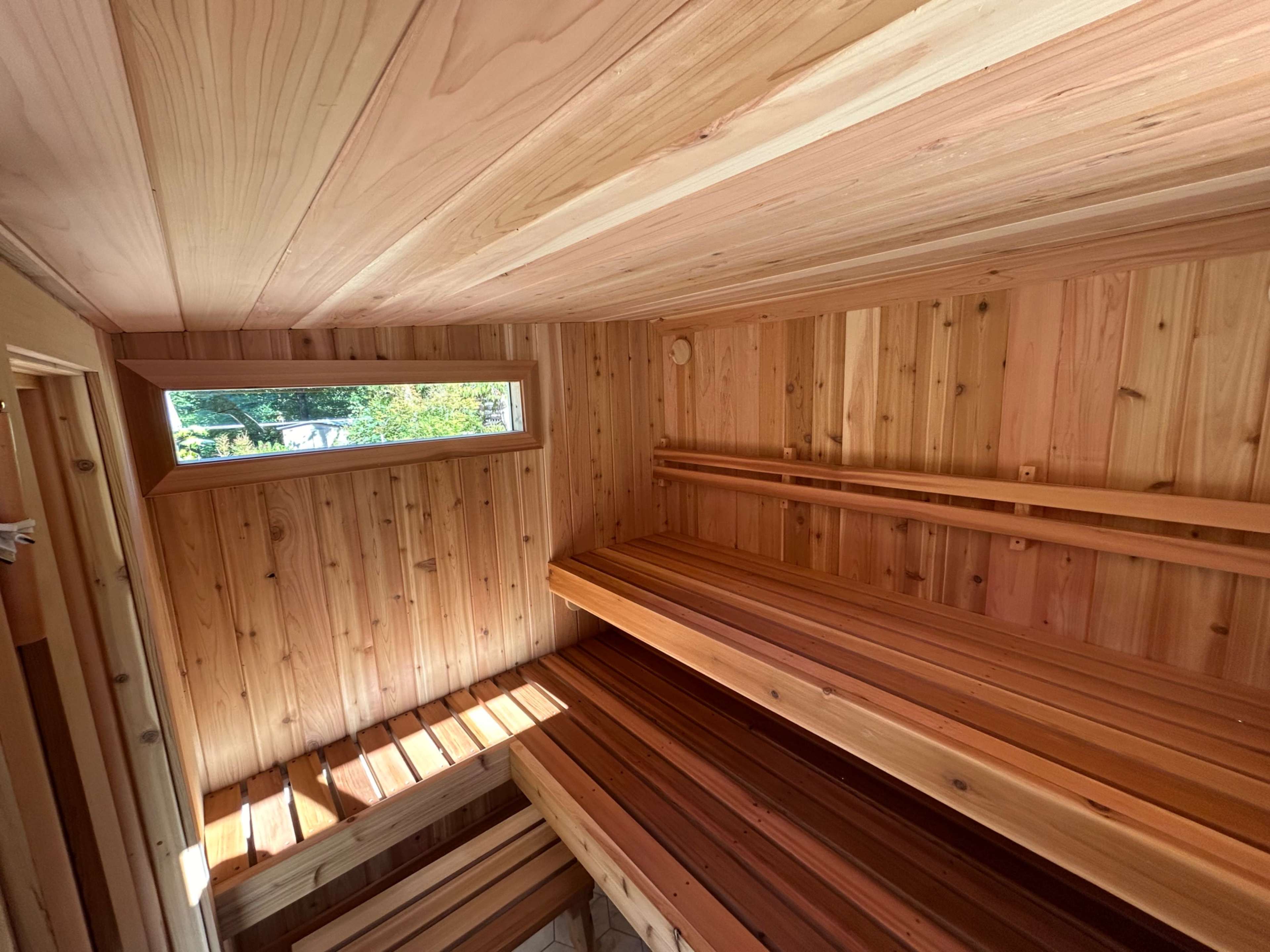 The interior of a wooden sauna features cedar benches and walls, with a window allowing natural light to enter the space.