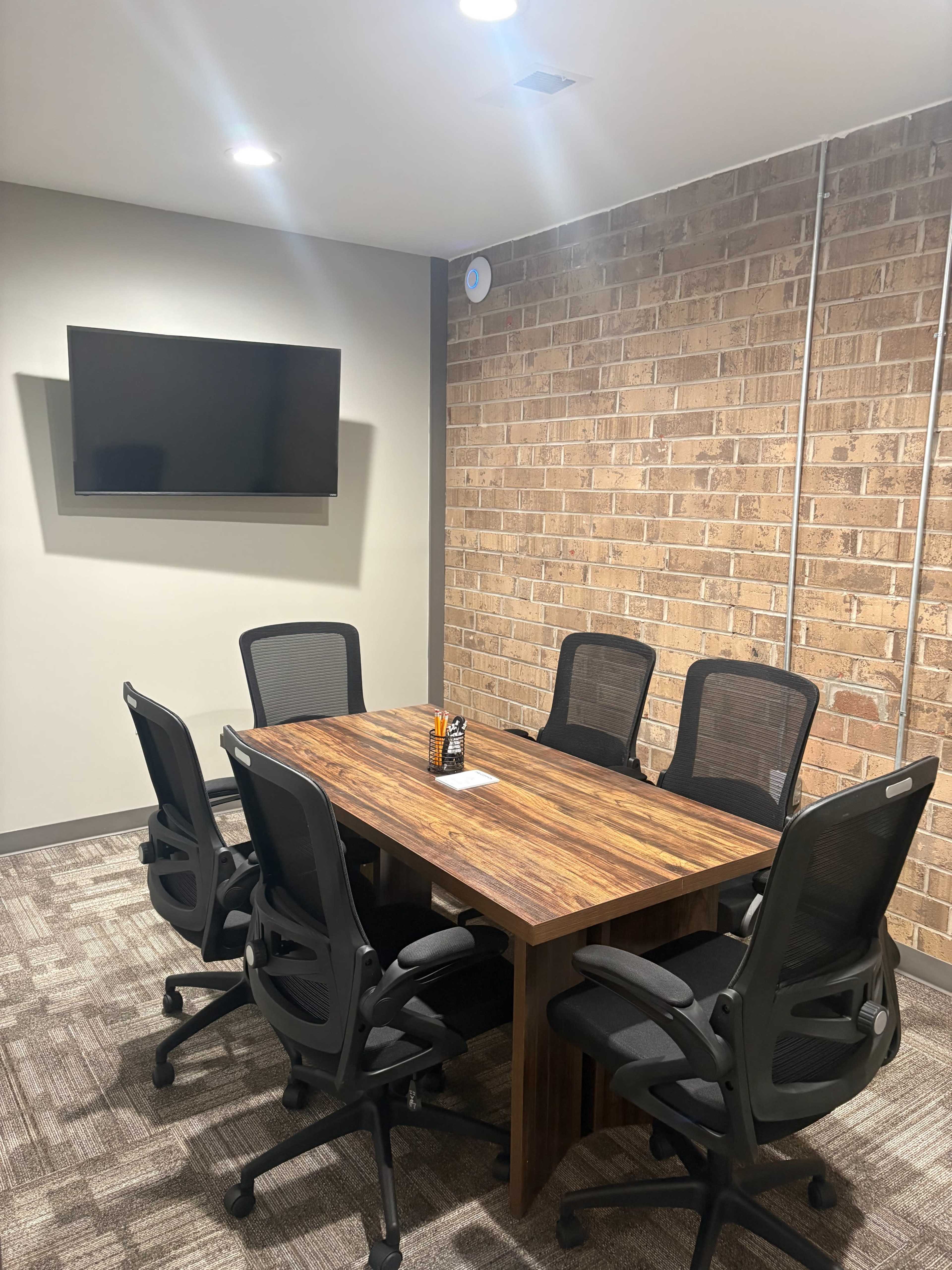 A conference room features a wooden table surrounded by six black office chairs, with a television mounted on the wall and exposed brick in the background.