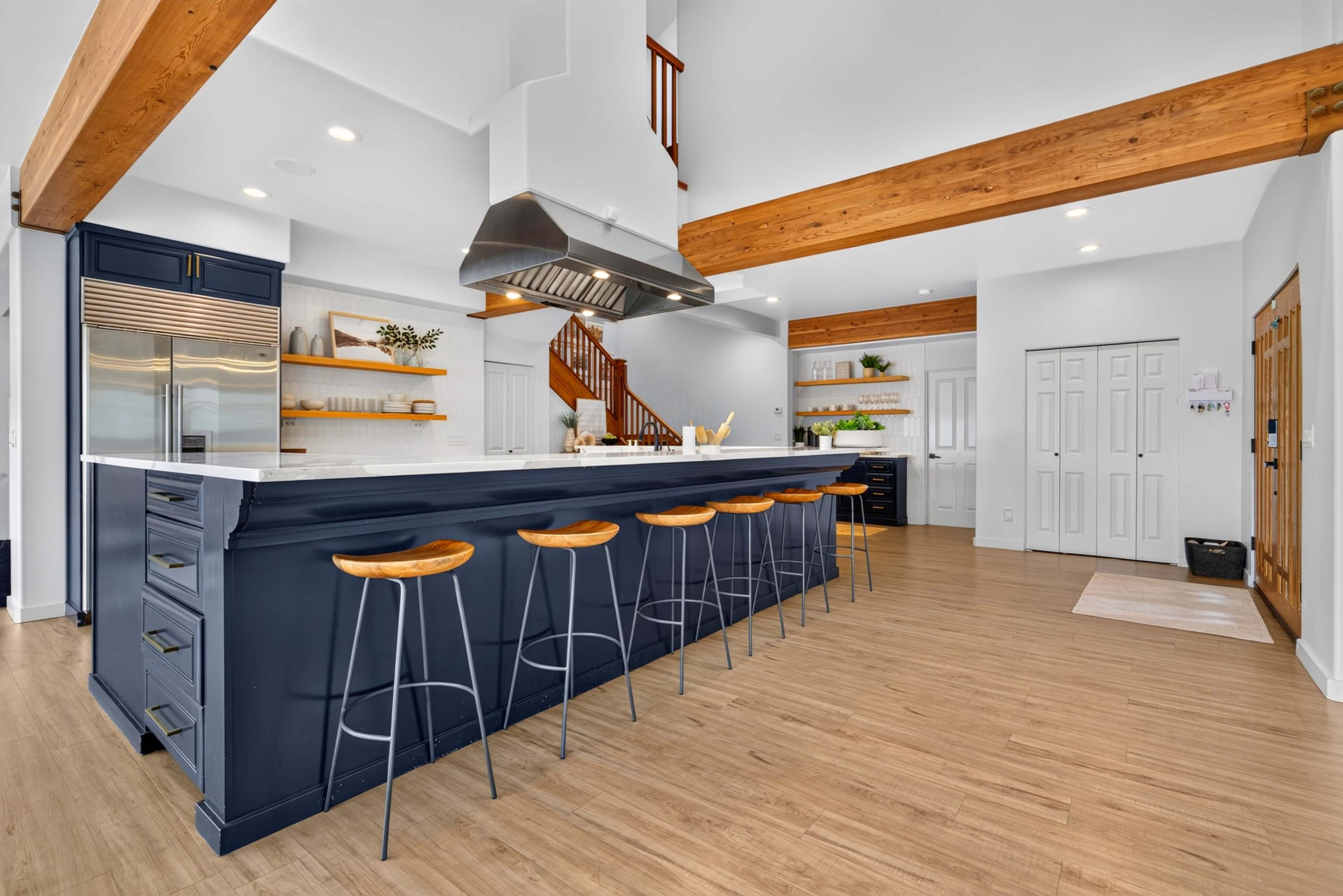 The image showcases a modern kitchen with a large island featuring bar stools, wooden beams overhead, and a staircase in the background.