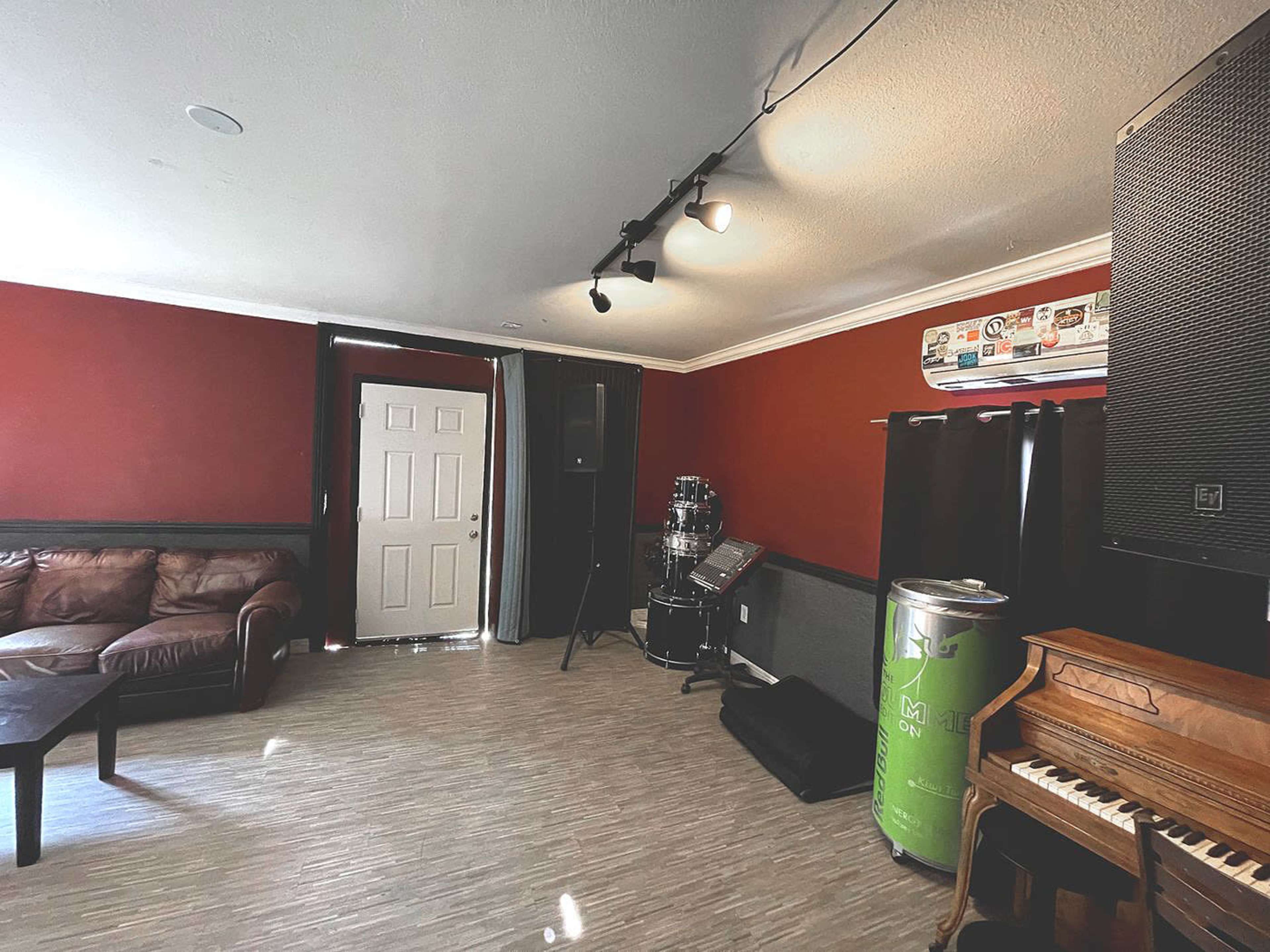 The image shows a music practice room featuring a brown couch, a door, a drum set, a speaker, and a piano, all against a red wall.