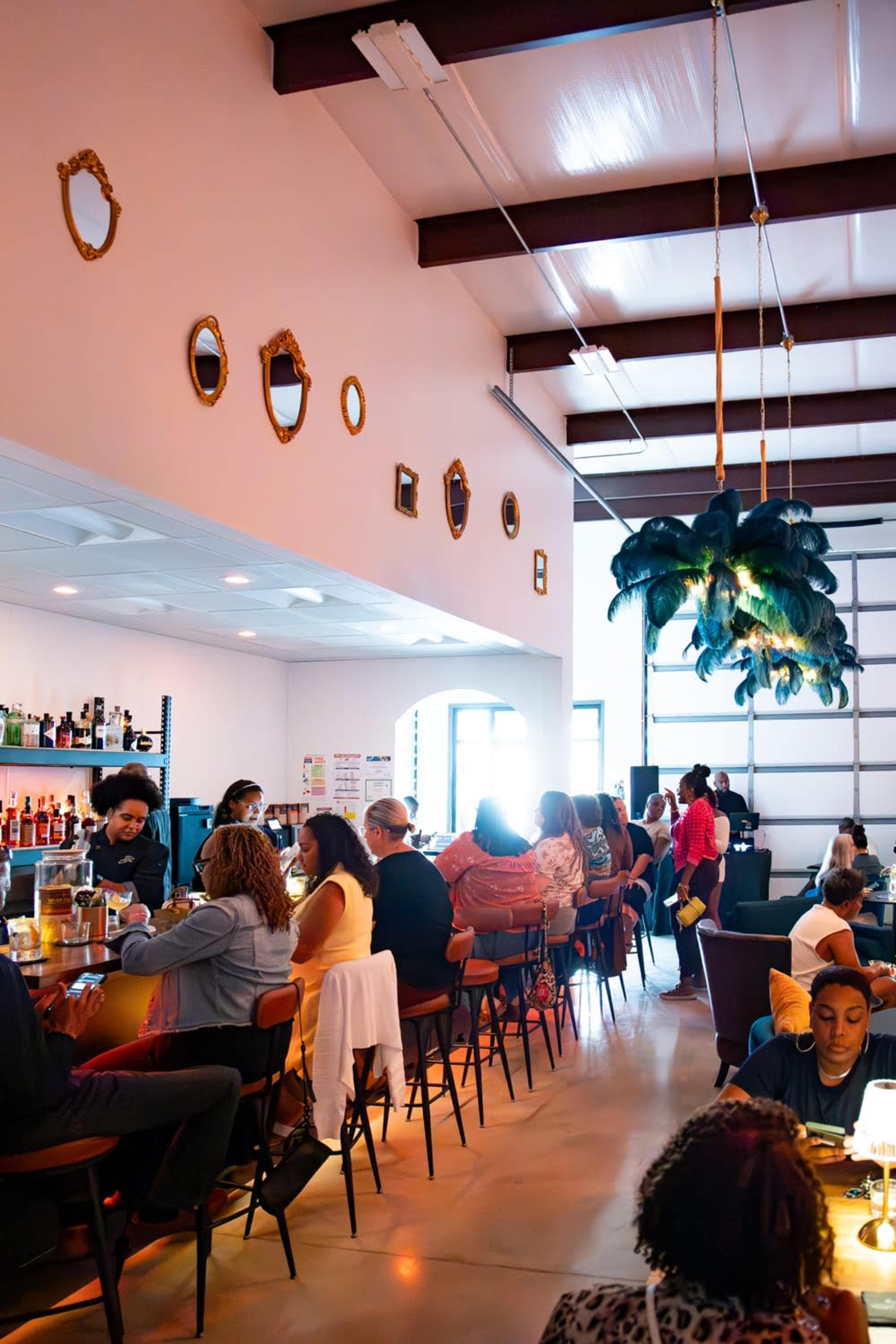 The image shows a lively bar scene inside a modern establishment, with patrons seated at a long counter and tables, as well as decorative mirrors and a large pendant light overhead.