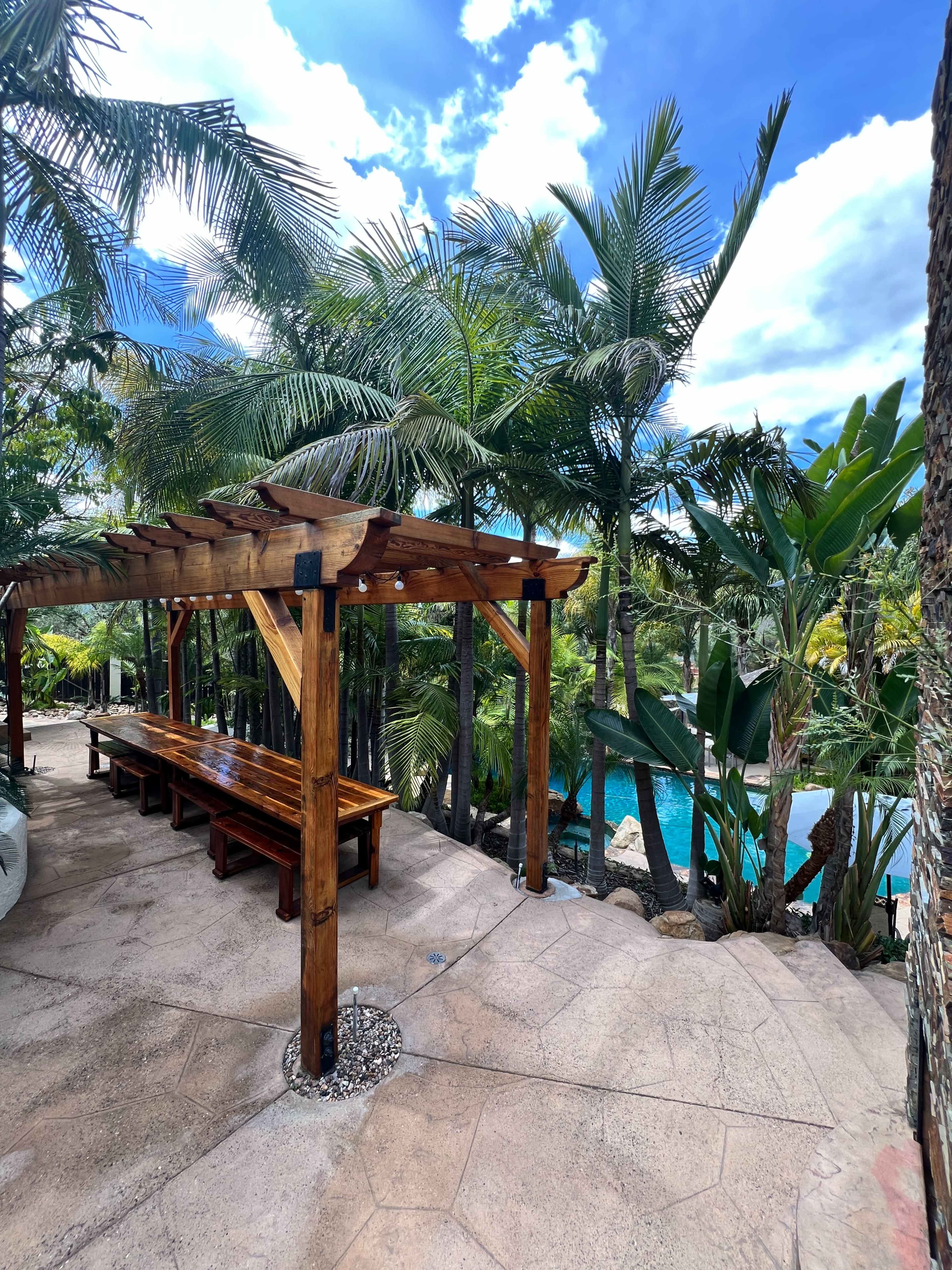 A wooden pergola with a long table is surrounded by lush palm trees and overlooks a blue pool.