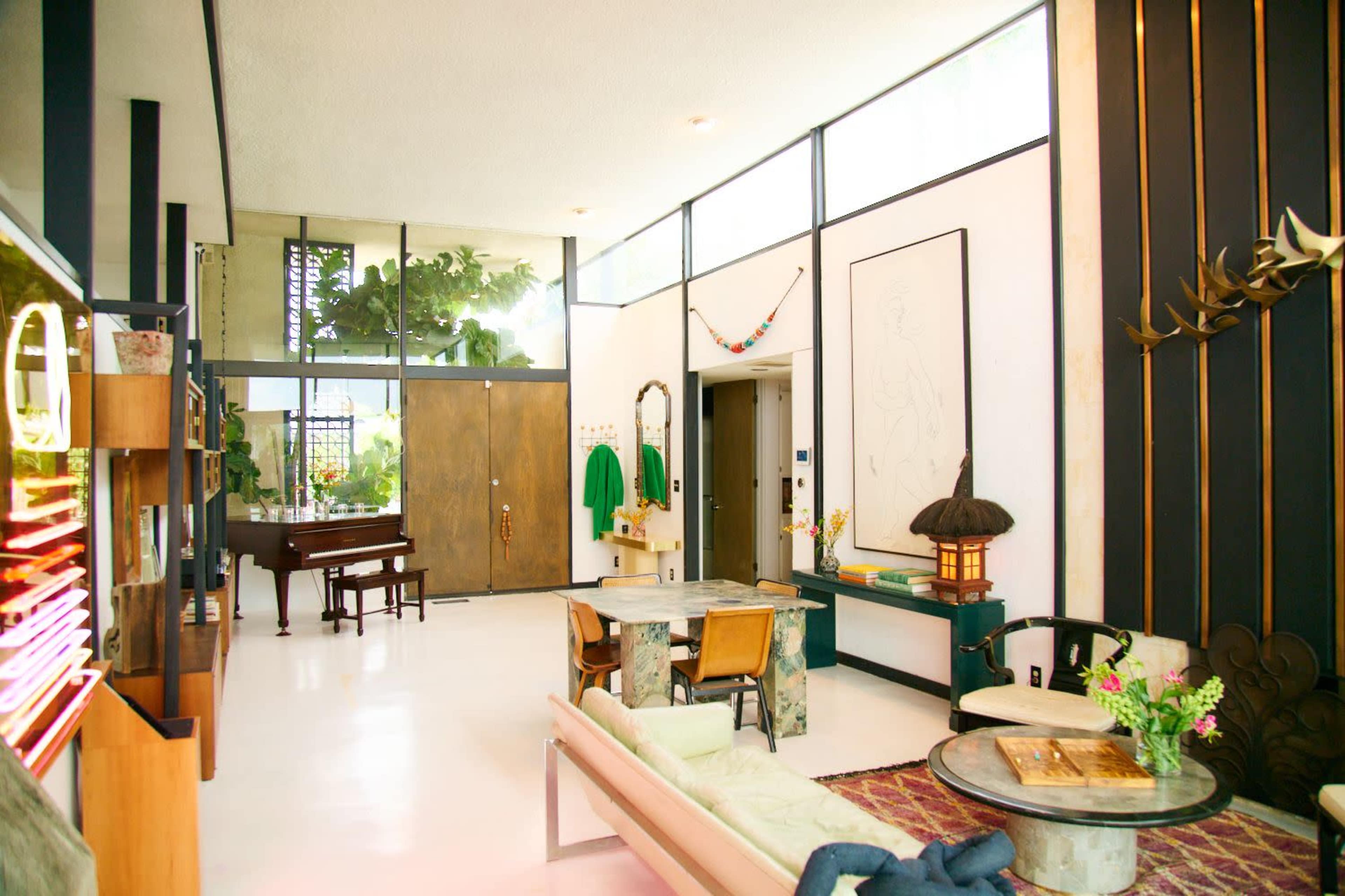 The image shows a mid-century modern living area featuring large windows, a grand piano, and minimalist furniture arranged around a coffee table.