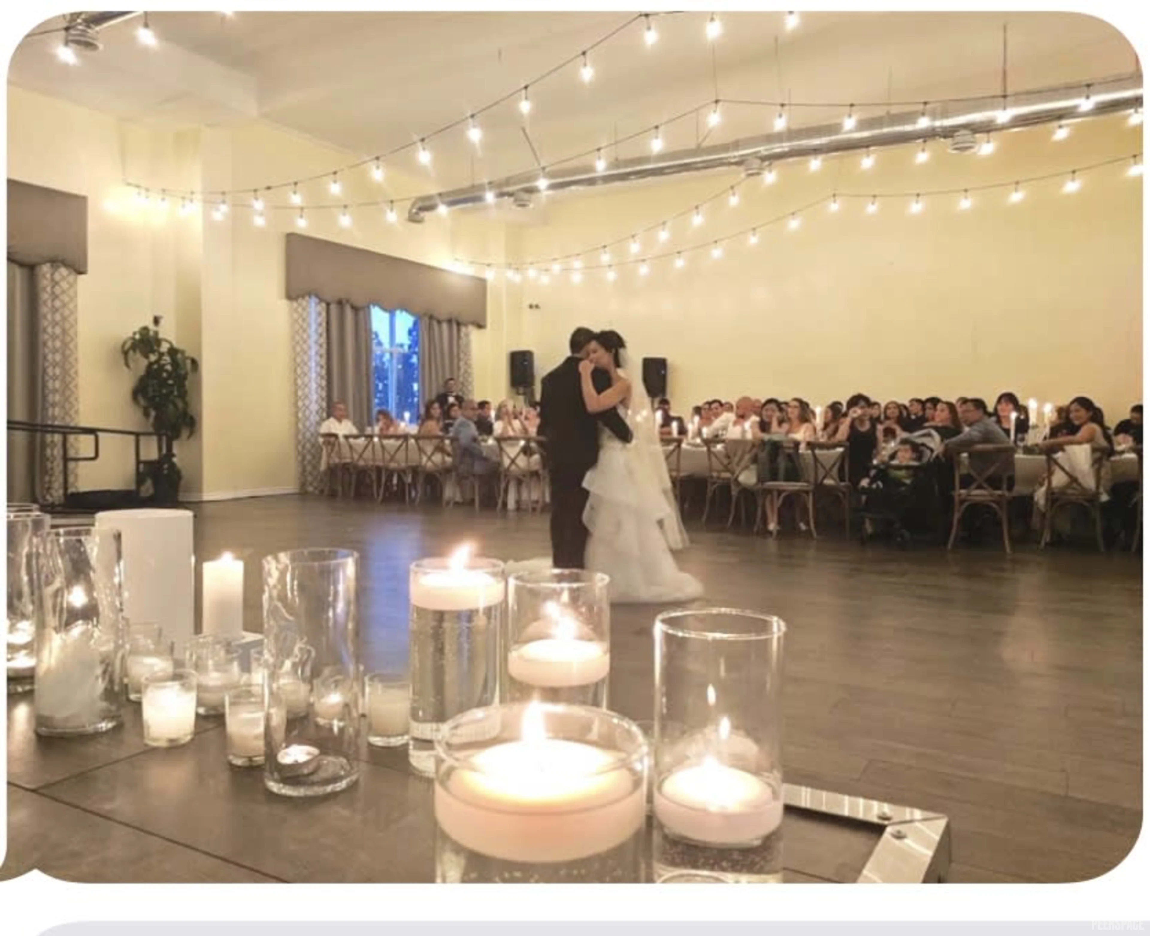 A couple dances together in a warmly lit reception hall while guests watch from tables adorned with candles.