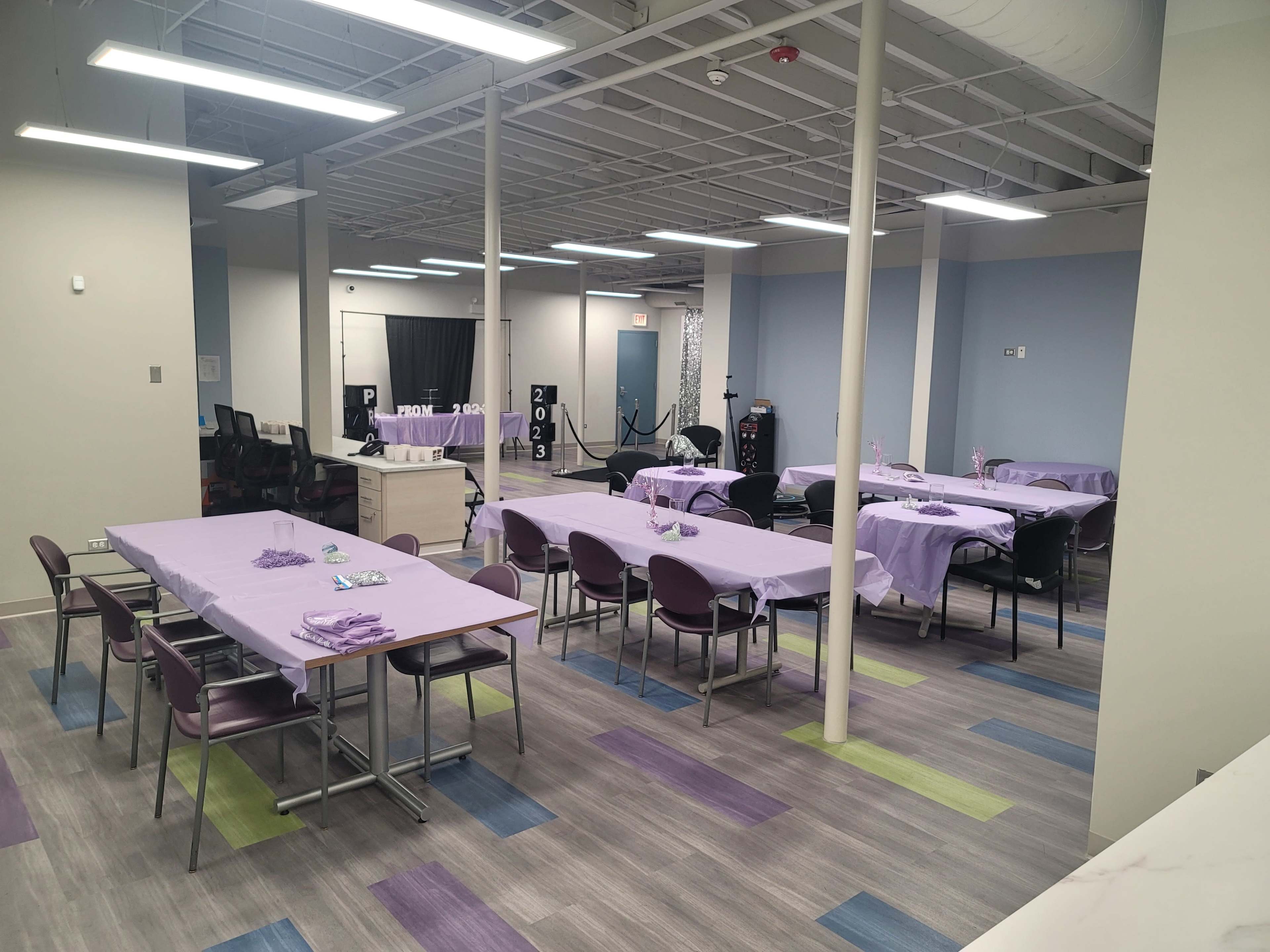 A spacious room is arranged with several tables covered in purple cloths, set up for an event, while chairs are positioned around them.