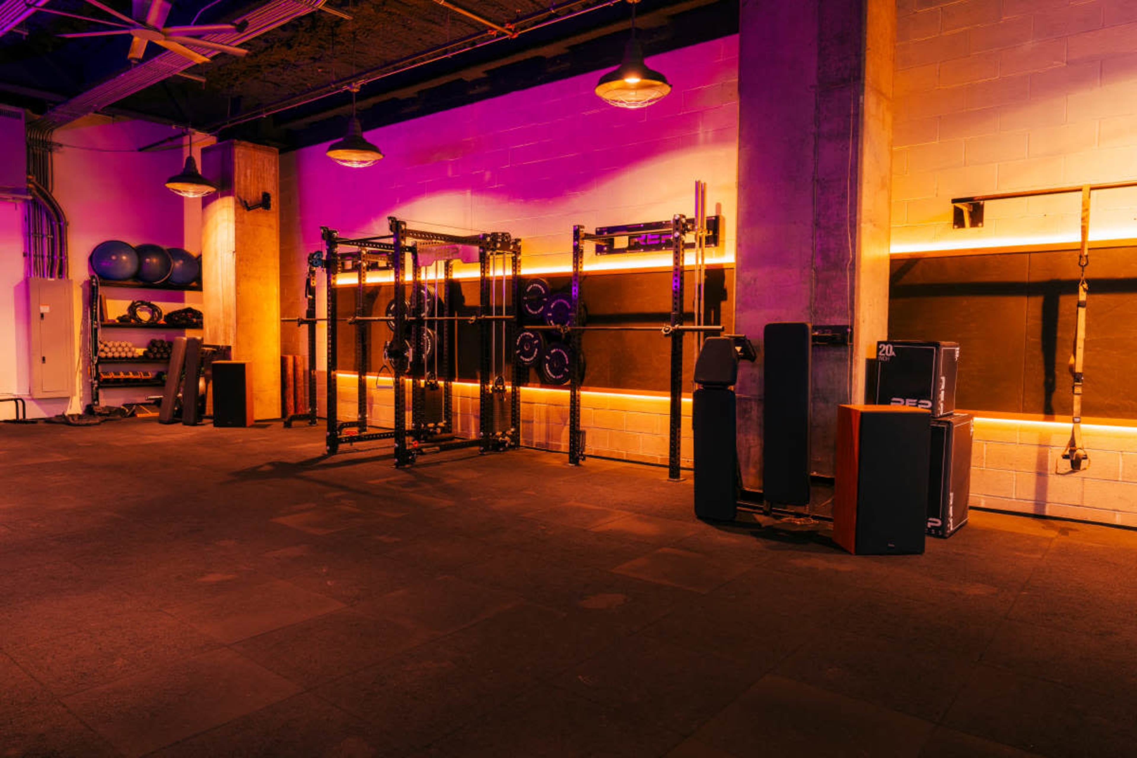 The image shows a gym interior with gym equipment, including weight racks and exercise balls, illuminated by purple and orange lighting.