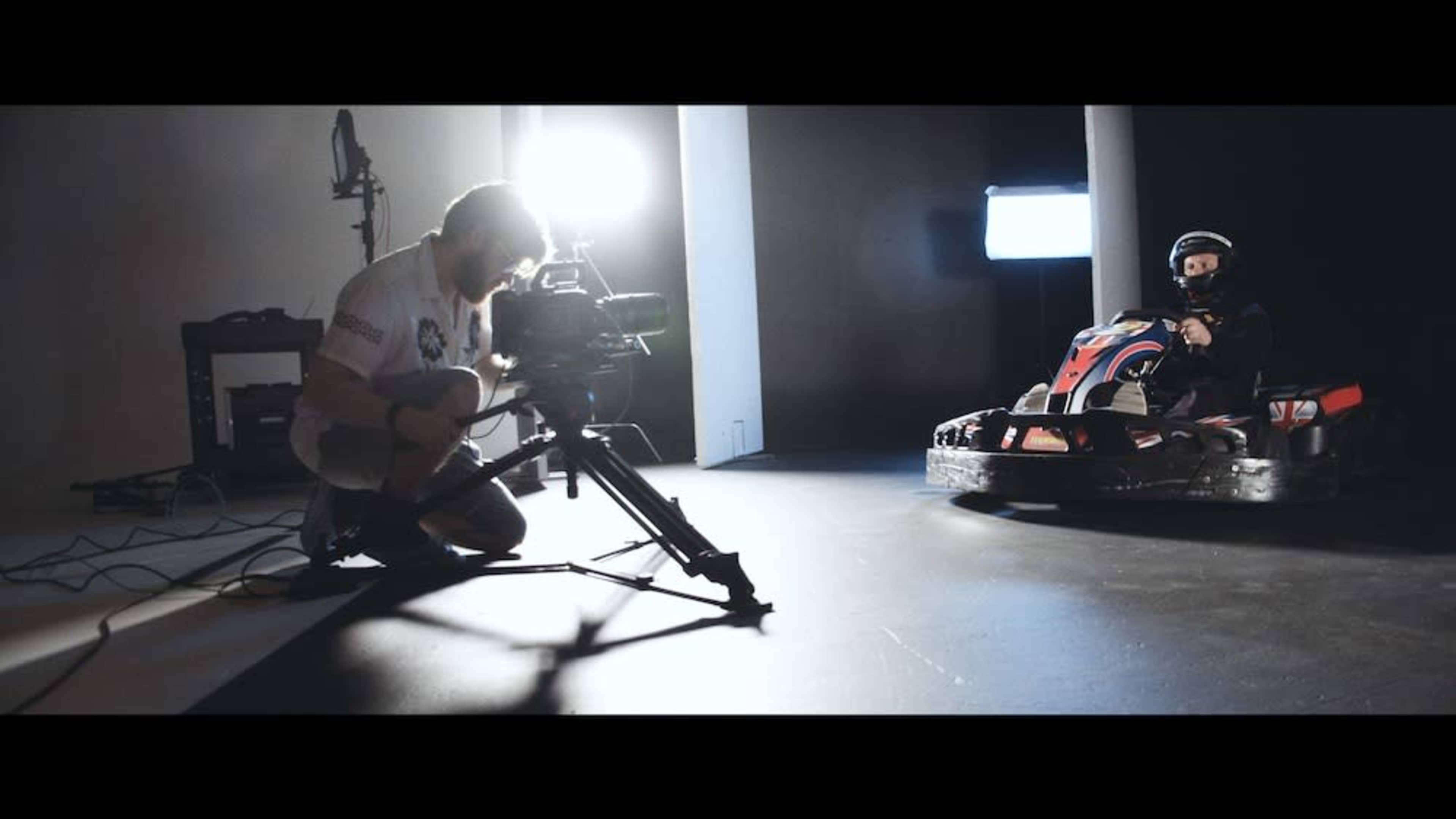 A camera operator sets up equipment while a child sits in a go-kart in a studio setting.