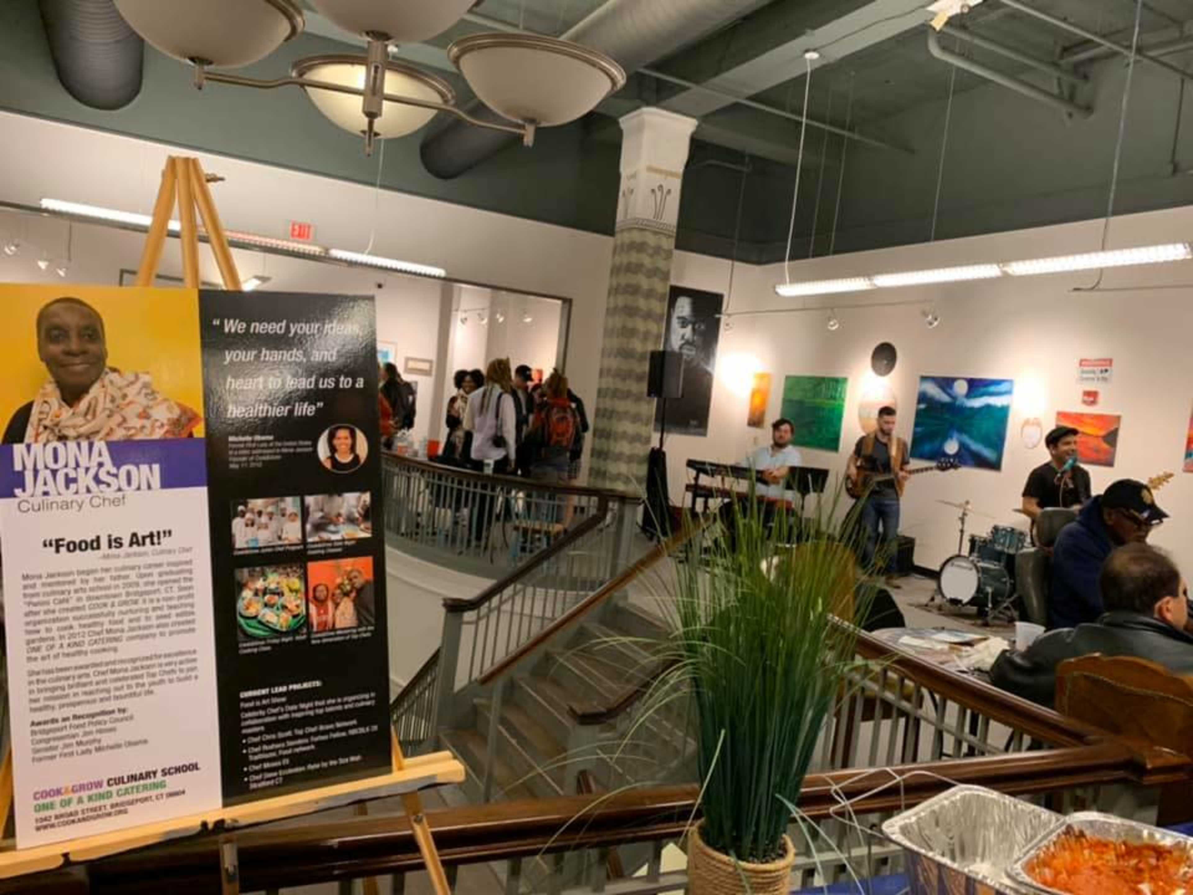 A crowd gathers in a gallery setting with artworks on the walls while a band performs and food is displayed in the foreground.