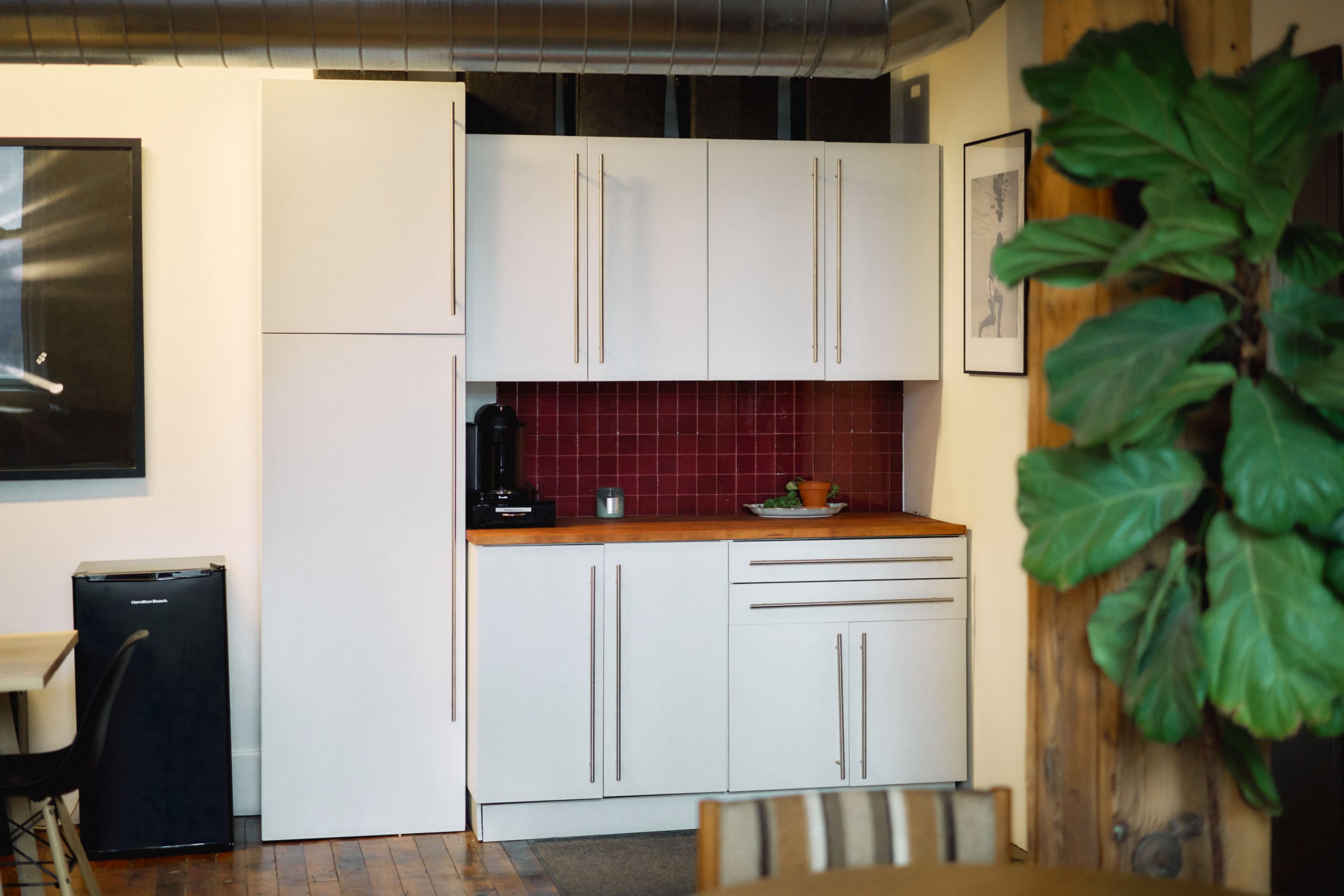 The image shows a modern kitchen corner featuring white cabinetry, a wooden countertop, and a dark red tiled backsplash, with a coffee machine and some kitchenware visible.