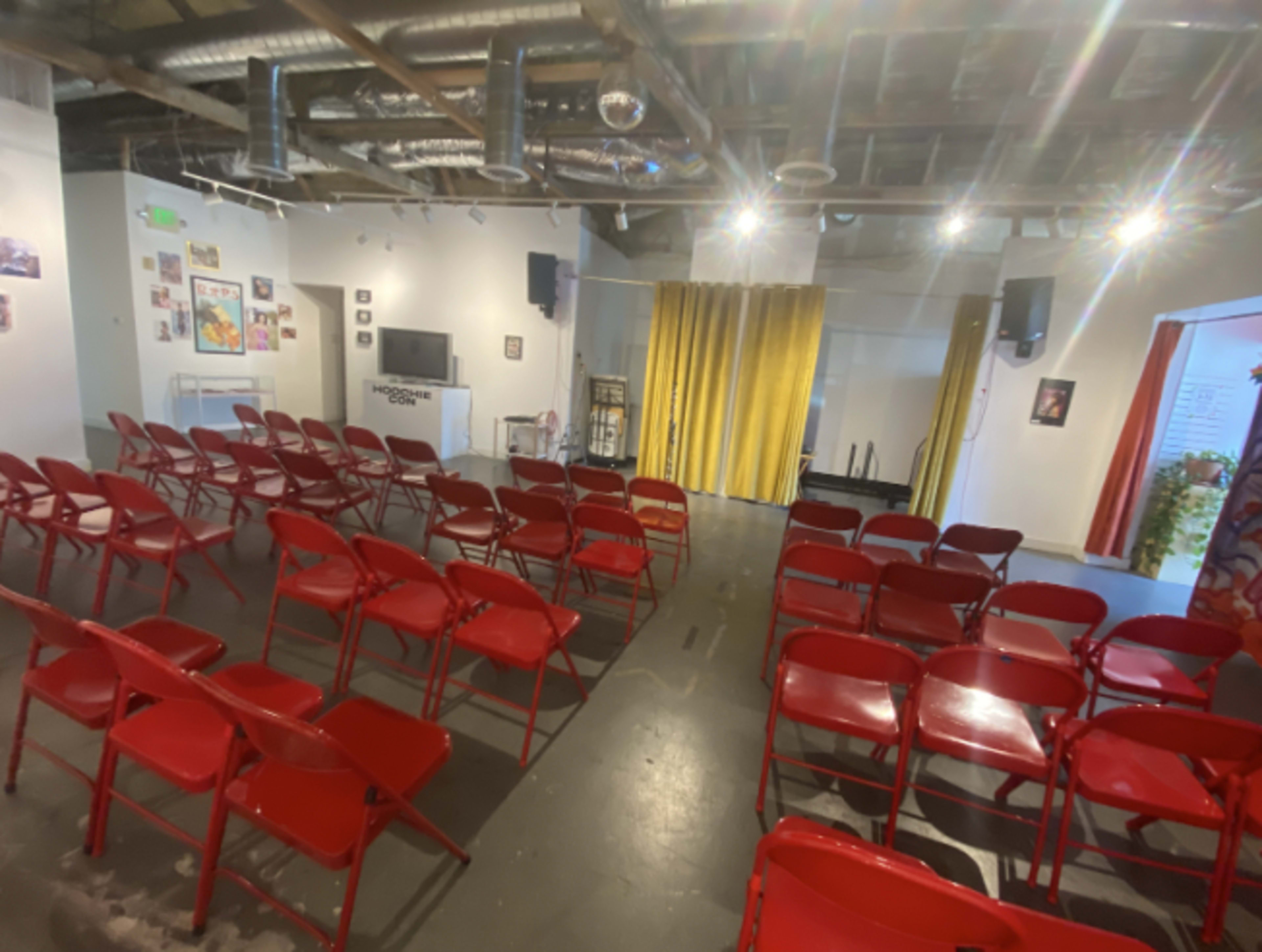 An empty audience area with red folding chairs arranged in rows facing a stage with yellow curtains and a television in the background.
