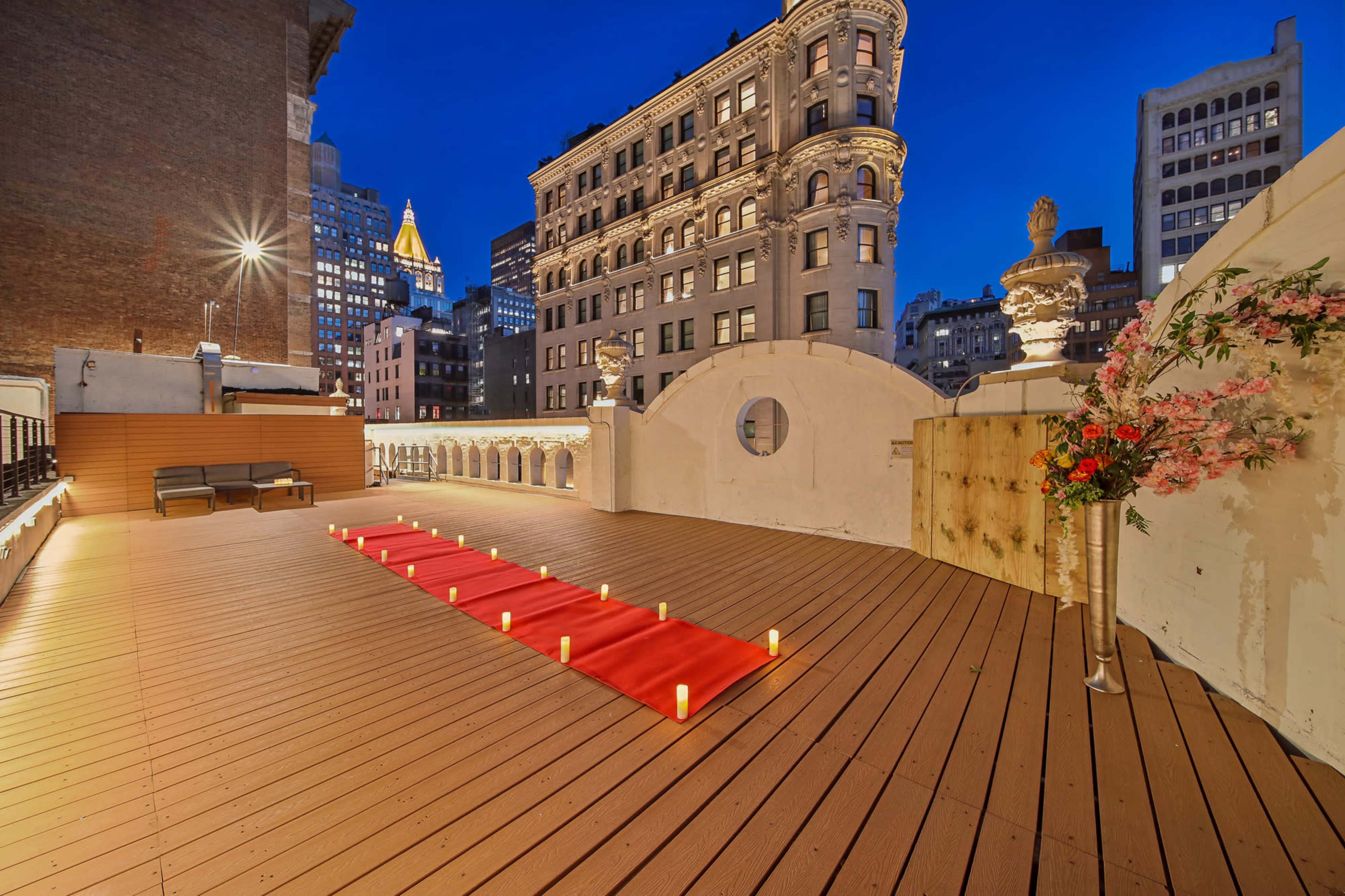 A rooftop terrace features a red carpet lined with candles, set against a backdrop of city buildings at night.
