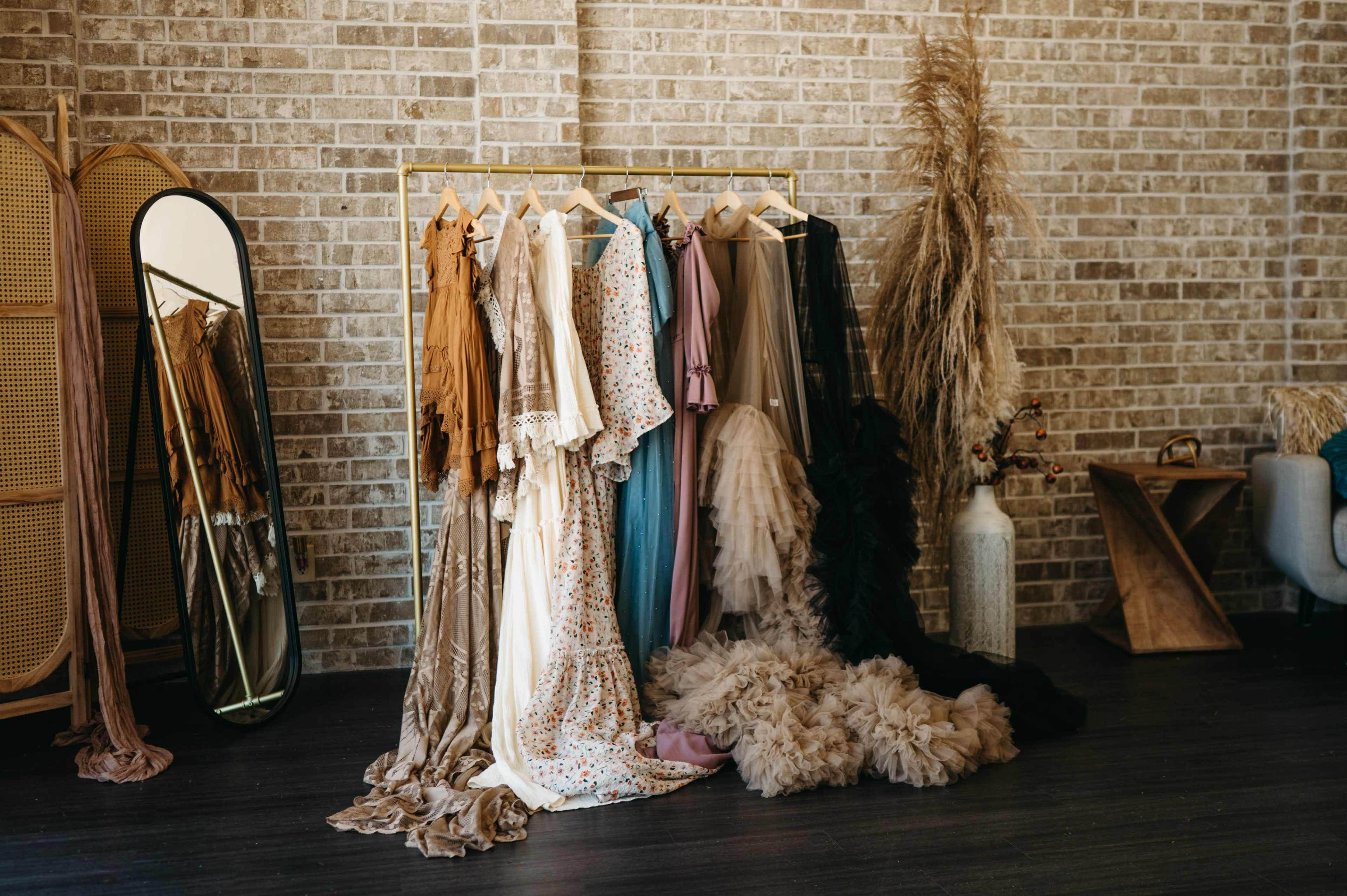 A variety of dresses in different colors and styles are hanging on a rack against a brick wall, accompanied by decorative elements like a mirror and dried plants.