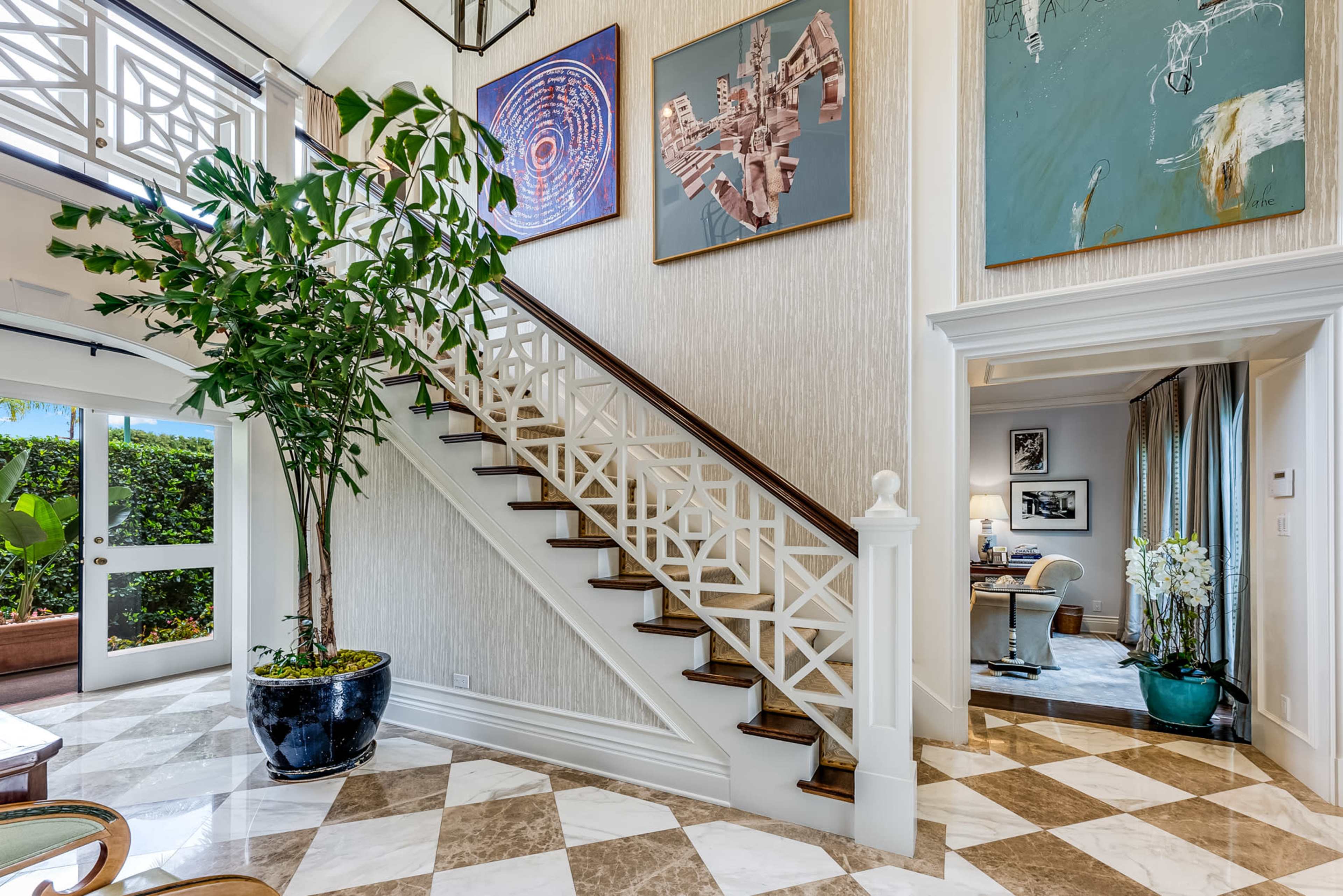 The image features a well-lit, spacious entryway with a staircase, decorative paintings on the walls, and a large potted plant.