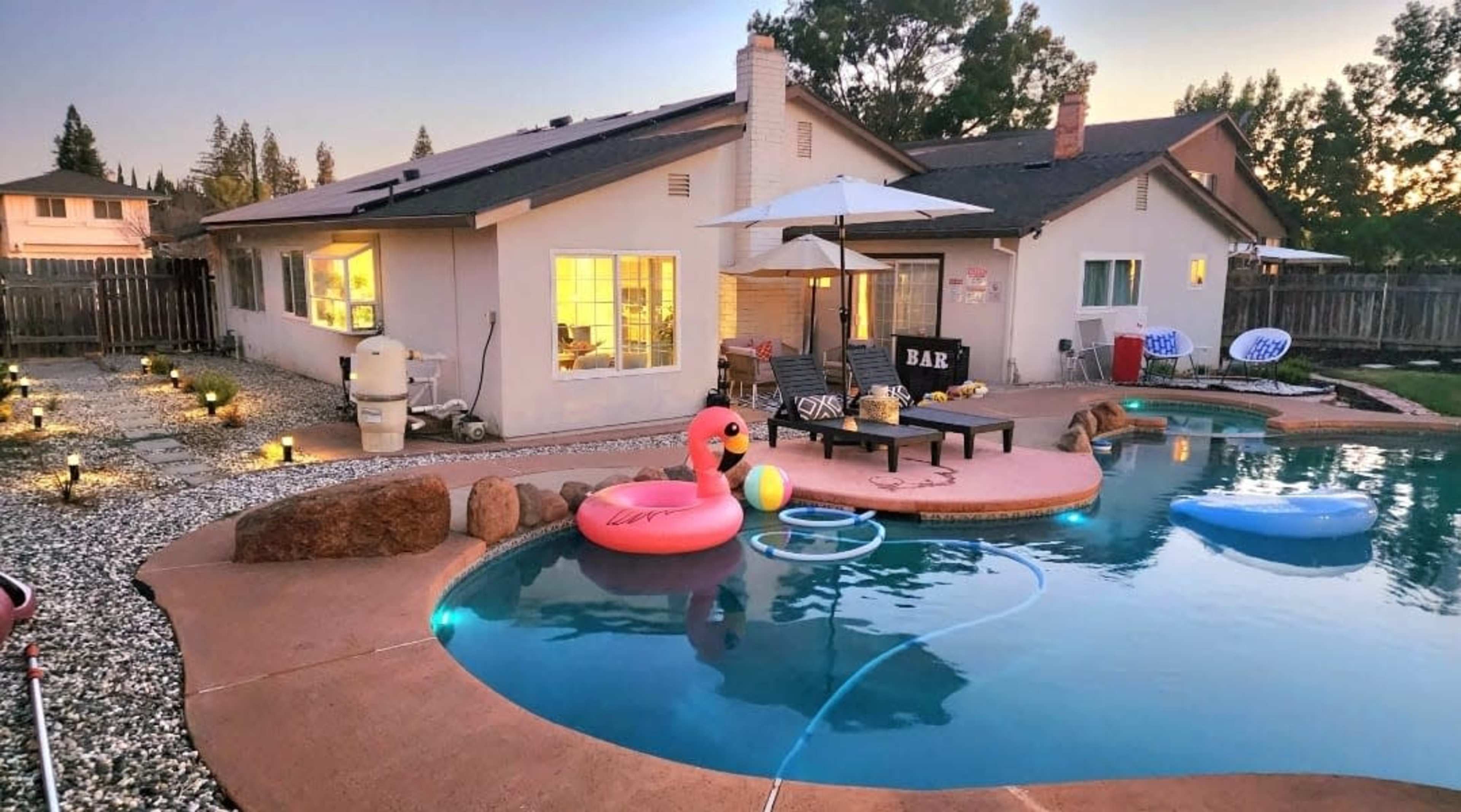 The image shows a backyard pool area with a swimming pool, colorful floats, a patio set, and a house in the background surrounded by landscaping.