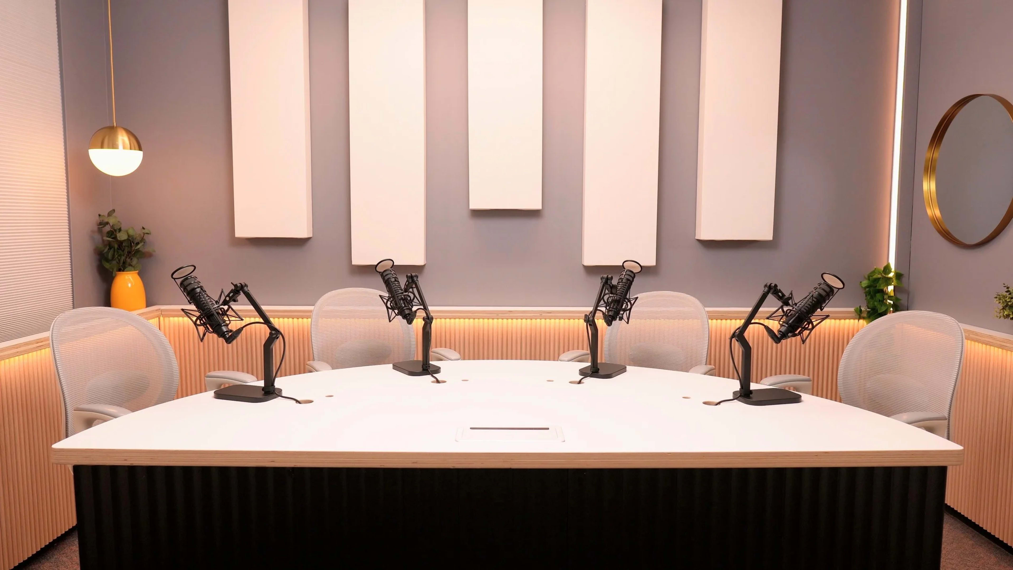 A modern podcast studio features a circular table with four microphone stands and white mesh chairs against a grey wall with soundproof panels.