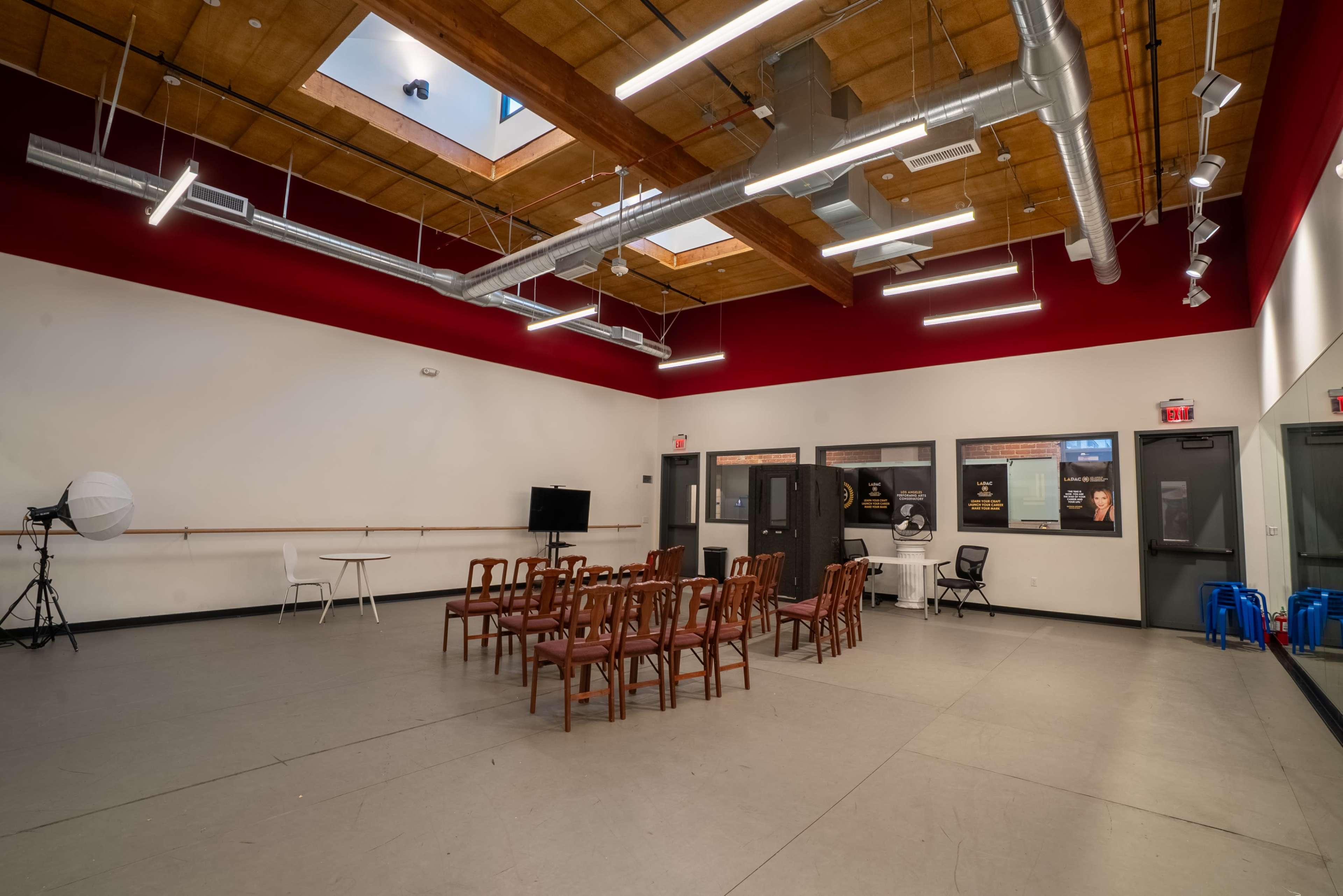 The image shows a rehearsal studio with rows of wooden chairs, a standing camera setup, and large windows allowing natural light.