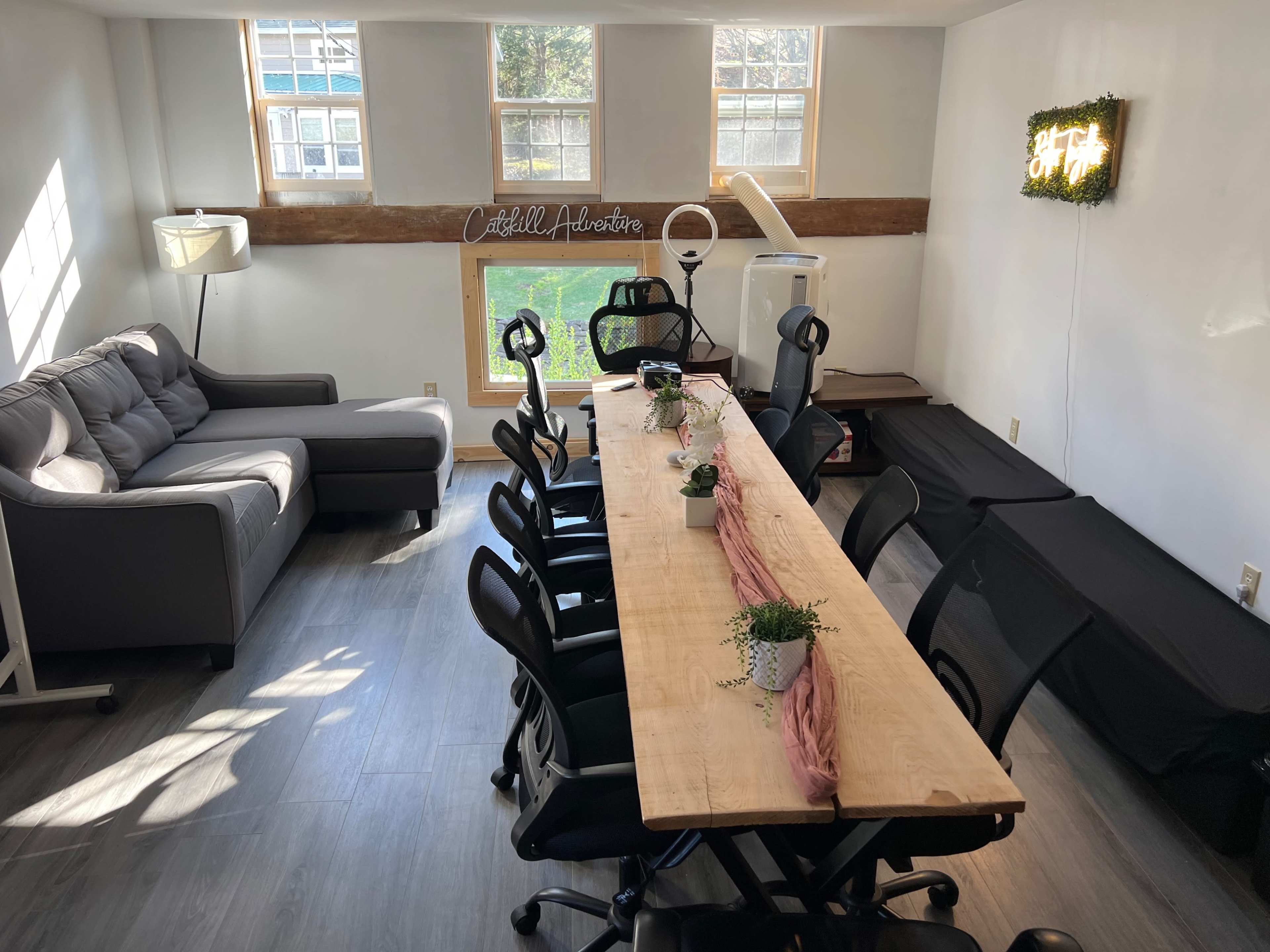 A well-lit room featuring a long wooden table with black chairs, a gray couch in the corner, and a window with a view of greenery outside.