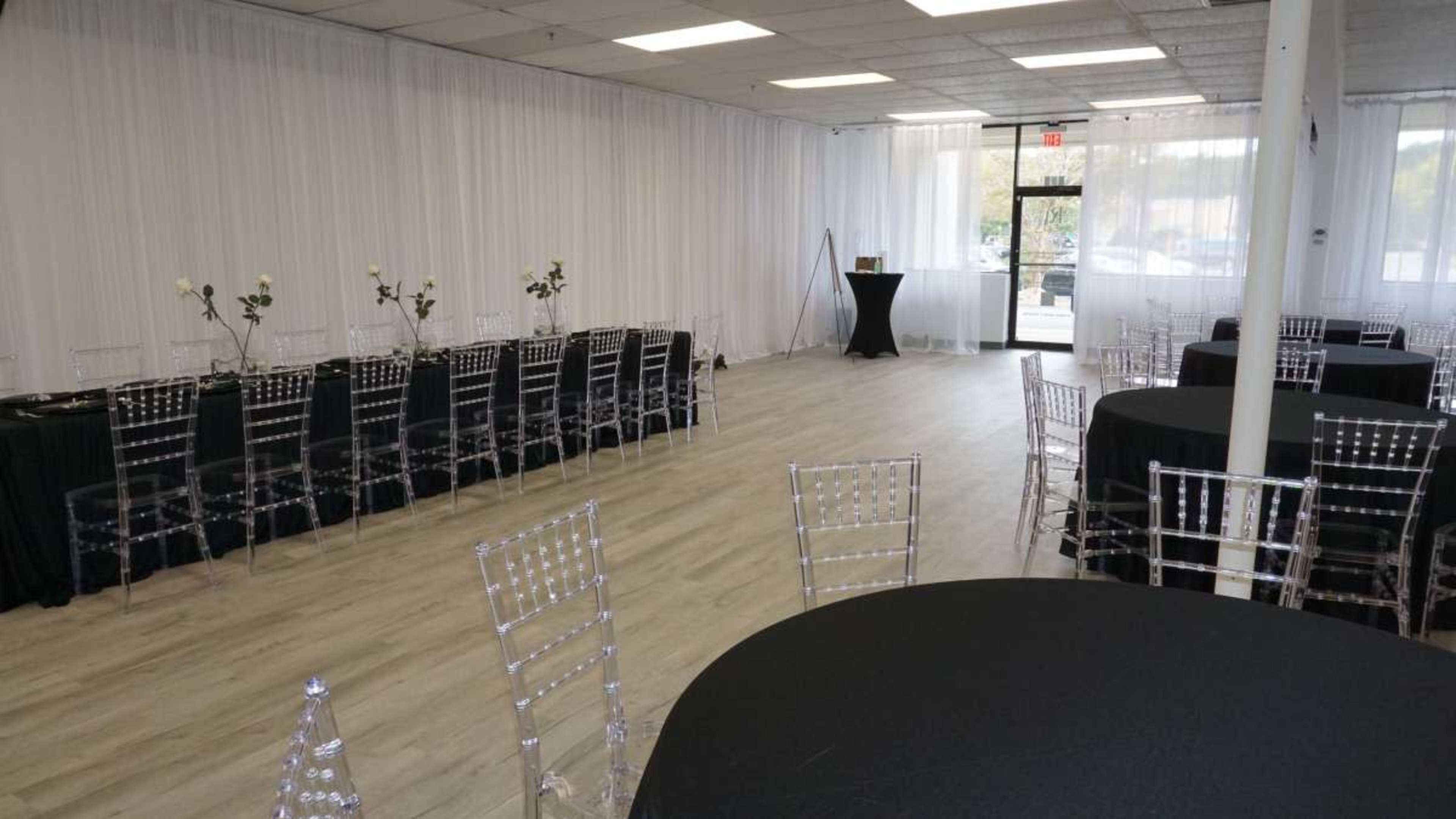 The image shows an event space with a light wooden floor, black tablecloths, clear acrylic chairs, and white drapery along the walls.