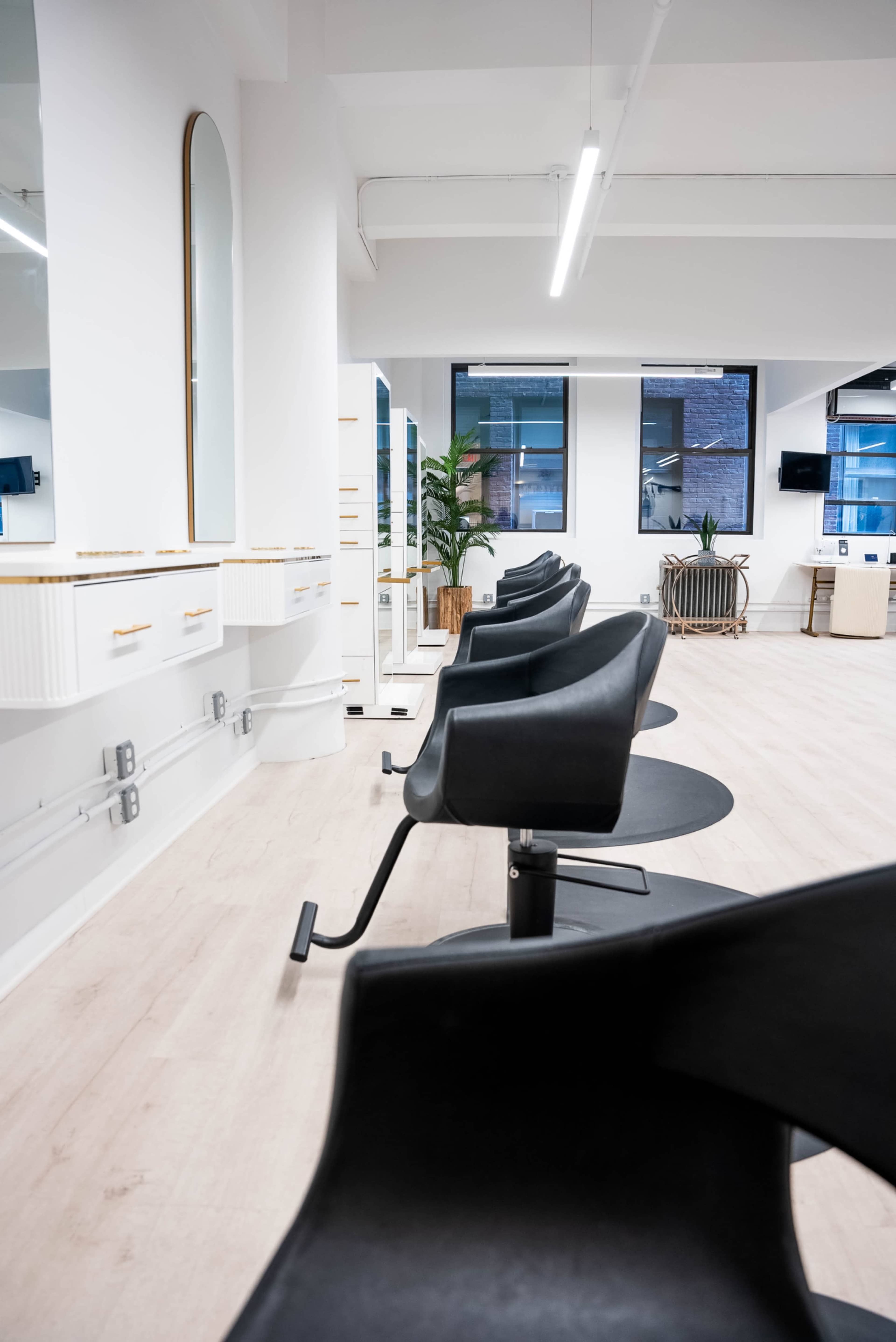 A modern hair salon with black styling chairs arranged in a row, illuminated by overhead lighting and featuring large mirrors along the walls.