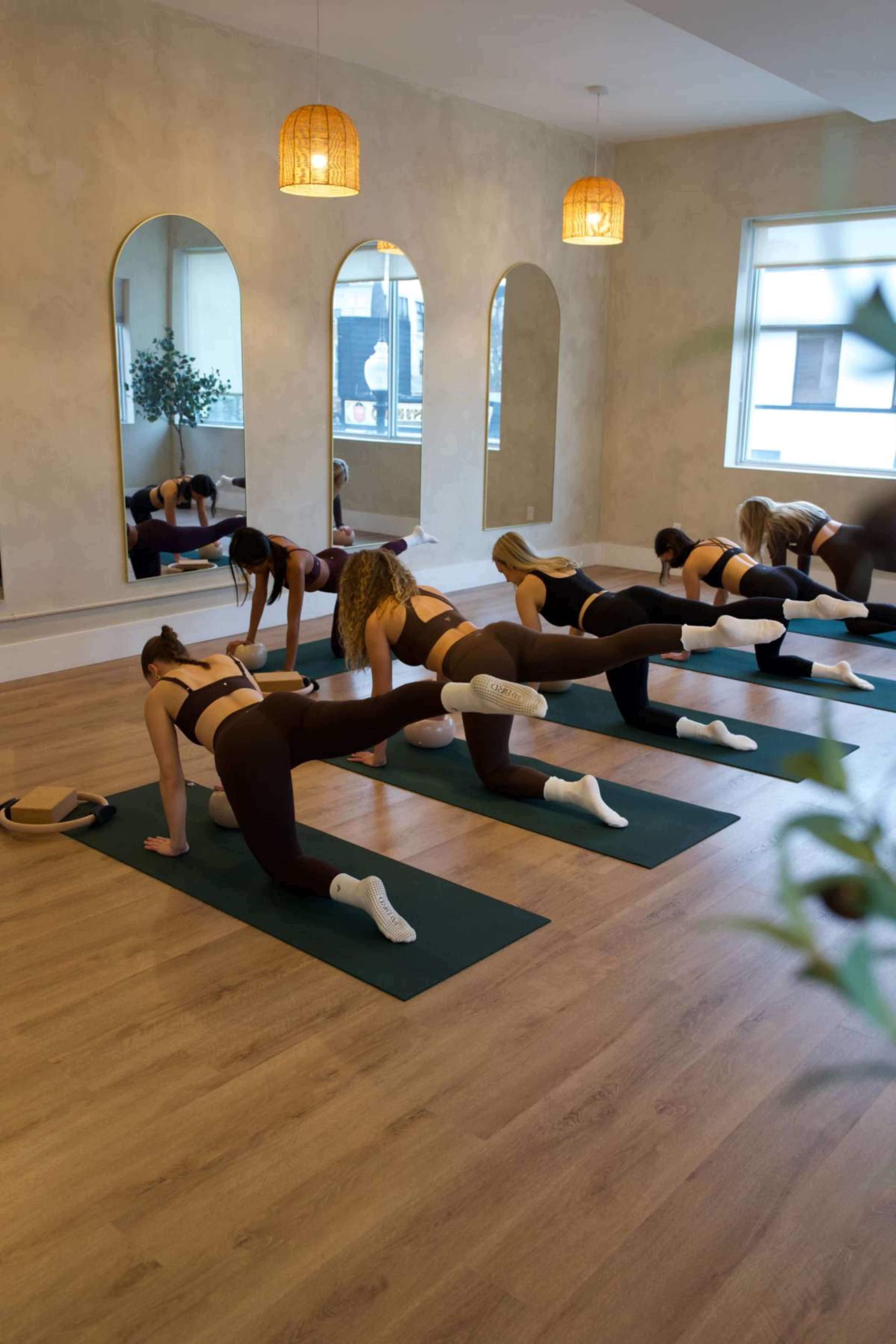 A group of people in workout attire is performing exercises on yoga mats in a well-lit studio with large mirrors and a neutral-colored wall.